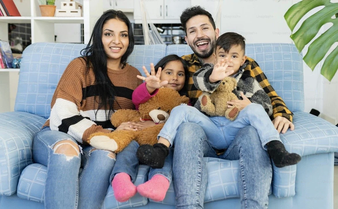 A family of five sitting on a blue couch in a living room, smiling and waving at the camera, holding teddy bears. The group includes two women, a man, and two children. The background features a bookshelf and a large green plant.