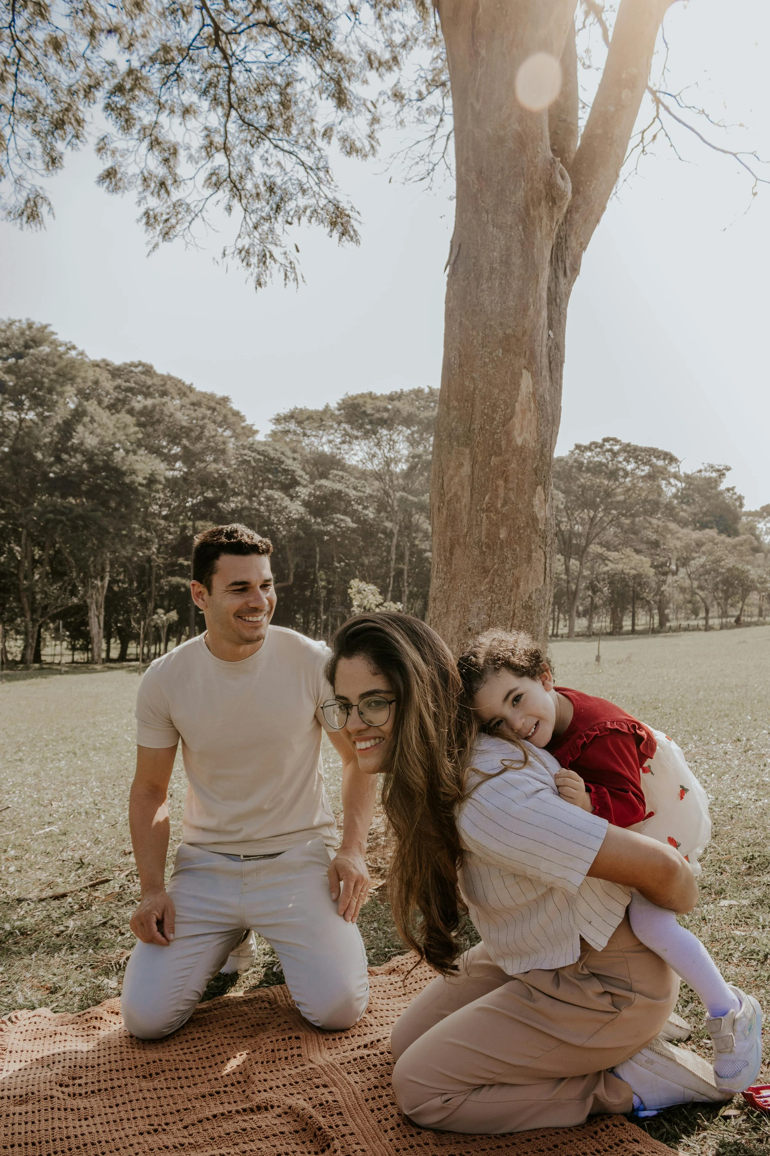 A family of four playing outdoors on a sunny day in a park with trees. The woman is kneeling on a blanket holding a young girl, and the man is kneeling next to them smiling. The young girl is resting her head on the woman, smiling at the camera. The family appears happy and enjoying their time together.