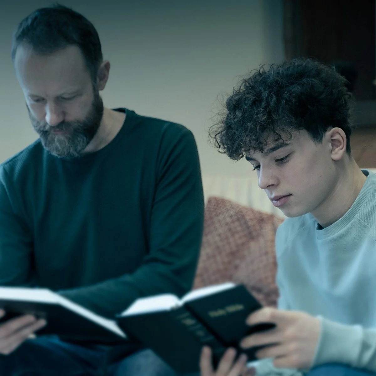 A man with a beard and a young man with curly hair sitting on a couch, reading a book together.