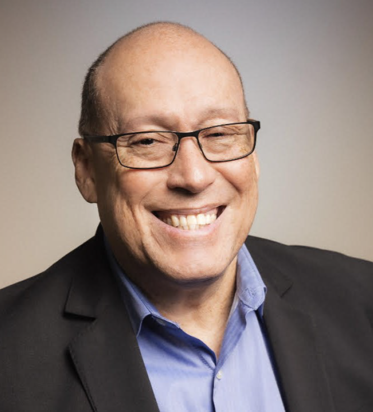 A smiling middle-aged man wearing glasses, a blue collared shirt, and a dark blazer.