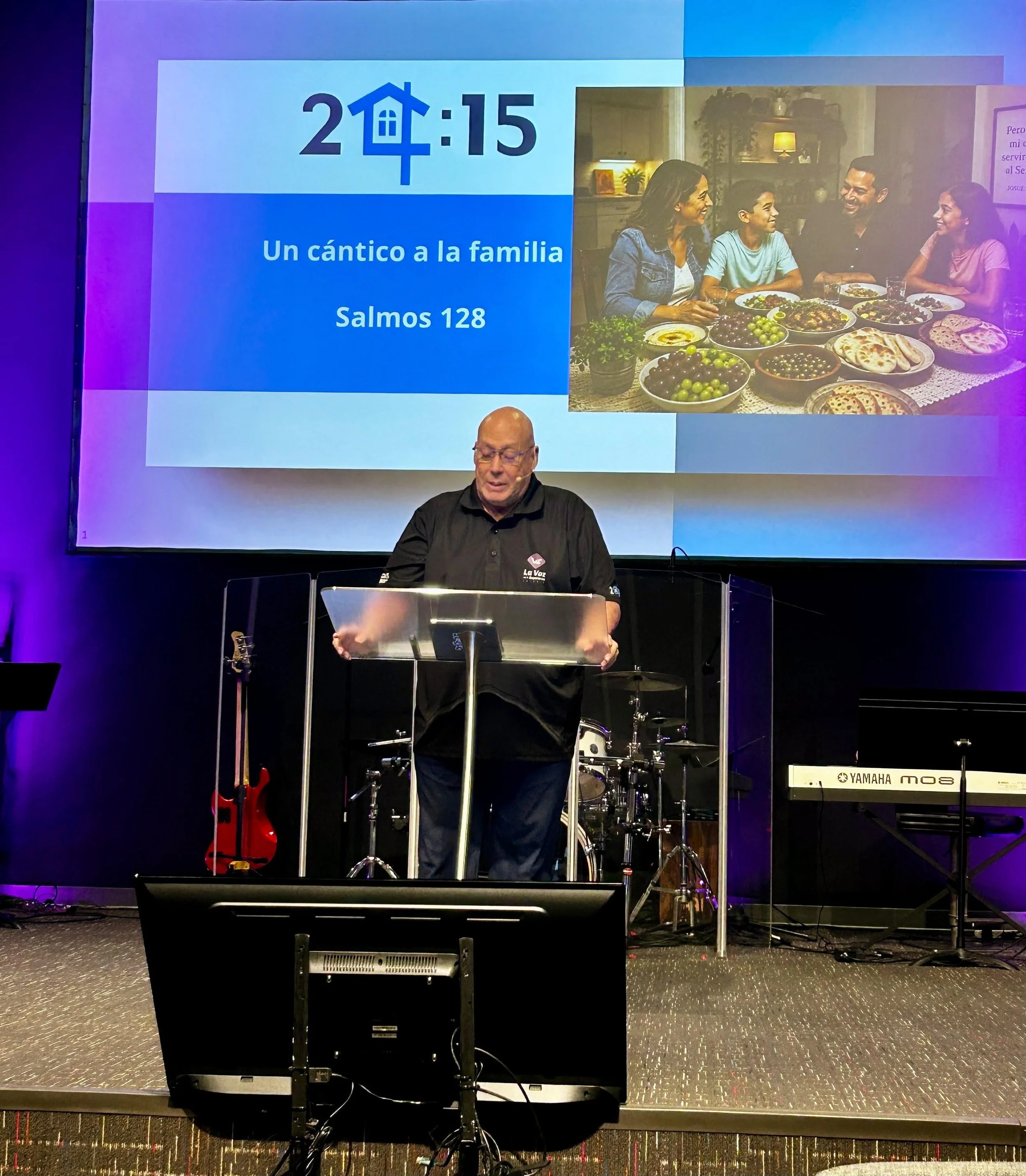 A man standing at a music stand with a musical instrument in front of him, behind him a large screen displaying a timer and the text 'Un cántico a la familia Salmos 128', and a photo of a family gathered around a table with food inside a room with wa
