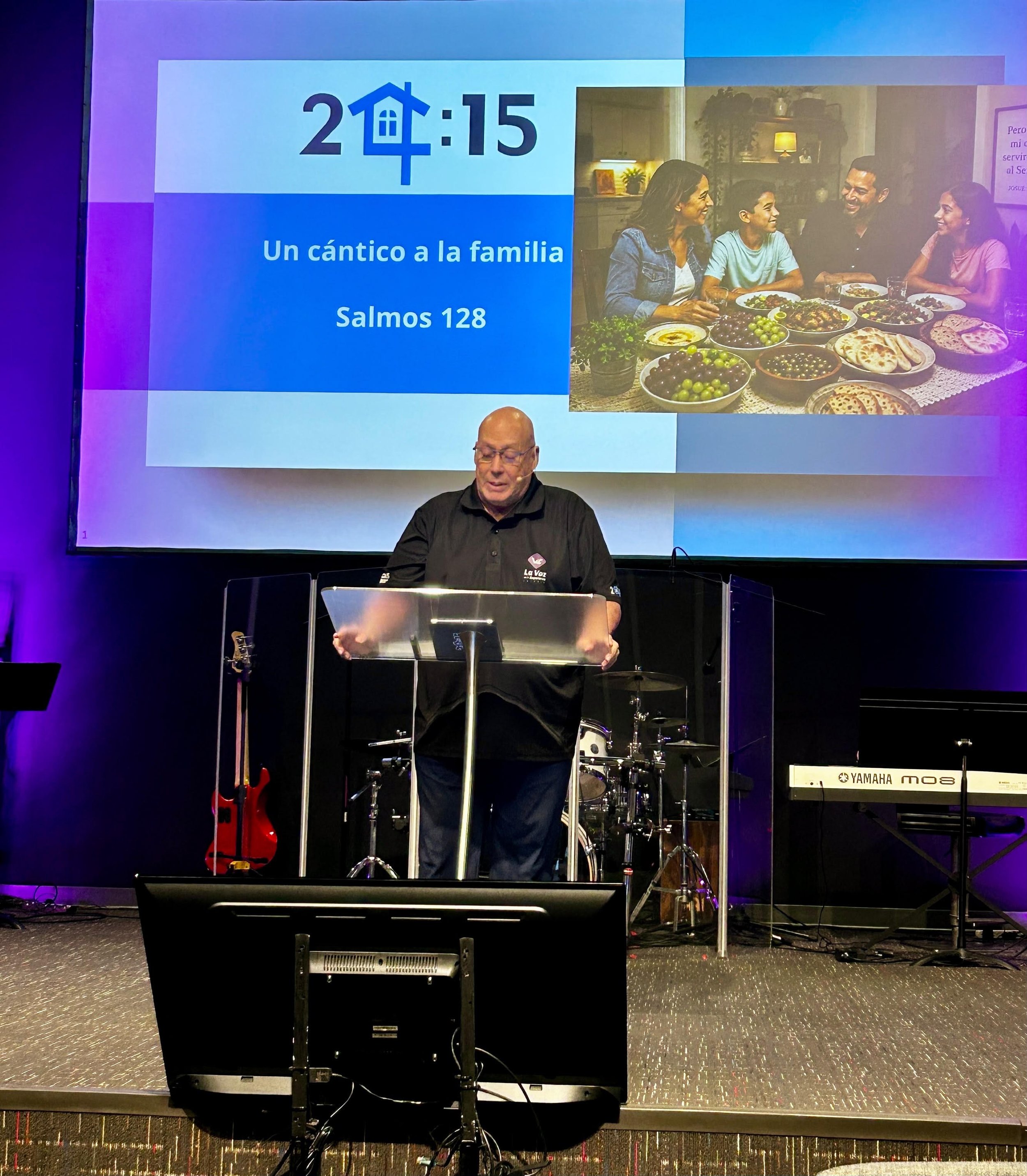 A man is standing behind a music stand on a stage, with a large screen behind him displaying the time 2:15, a house icon, and a Bible verse from Psalms 128. The screen also shows a photo of a family gathered around a table with food. The stage has mu