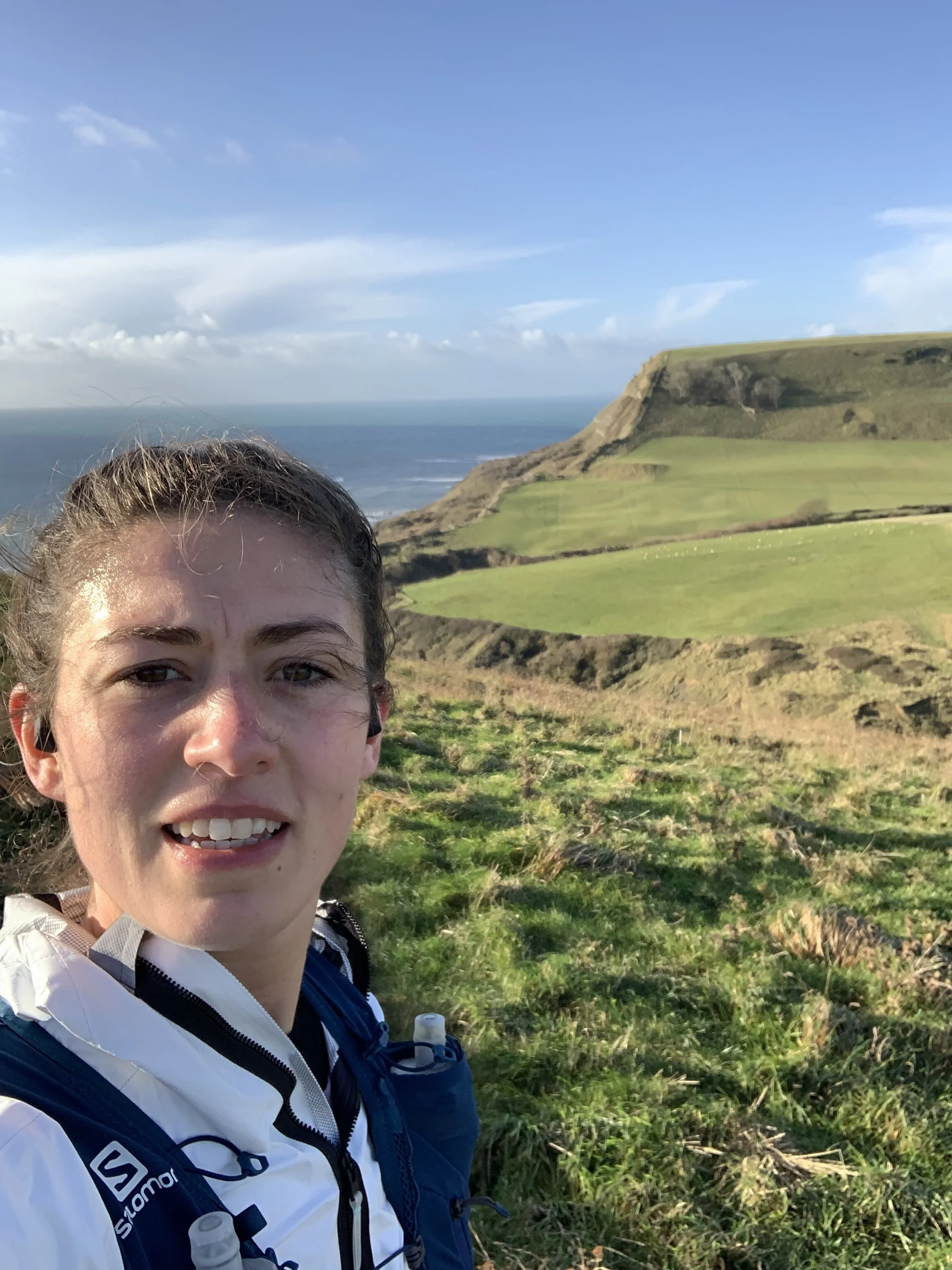 A young woman taking a selfie on a grassy hill with a scenic view of green cliffs, the ocean, and a blue sky with clouds in the background.