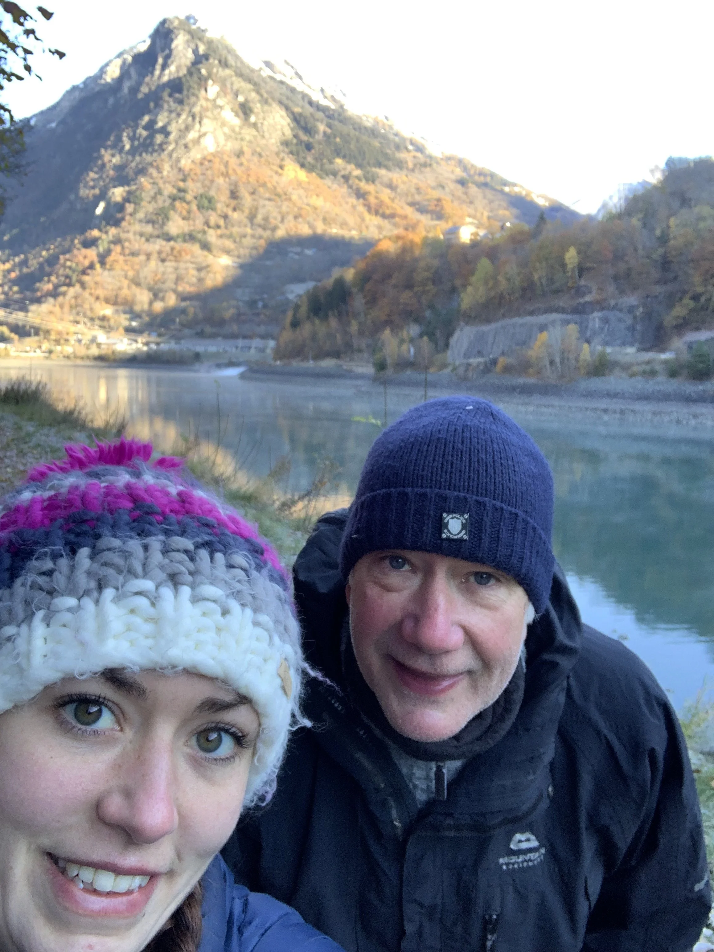 A smiling woman and man wearing winter hats and jackets taking a selfie outdoors near a river with mountains in the background.