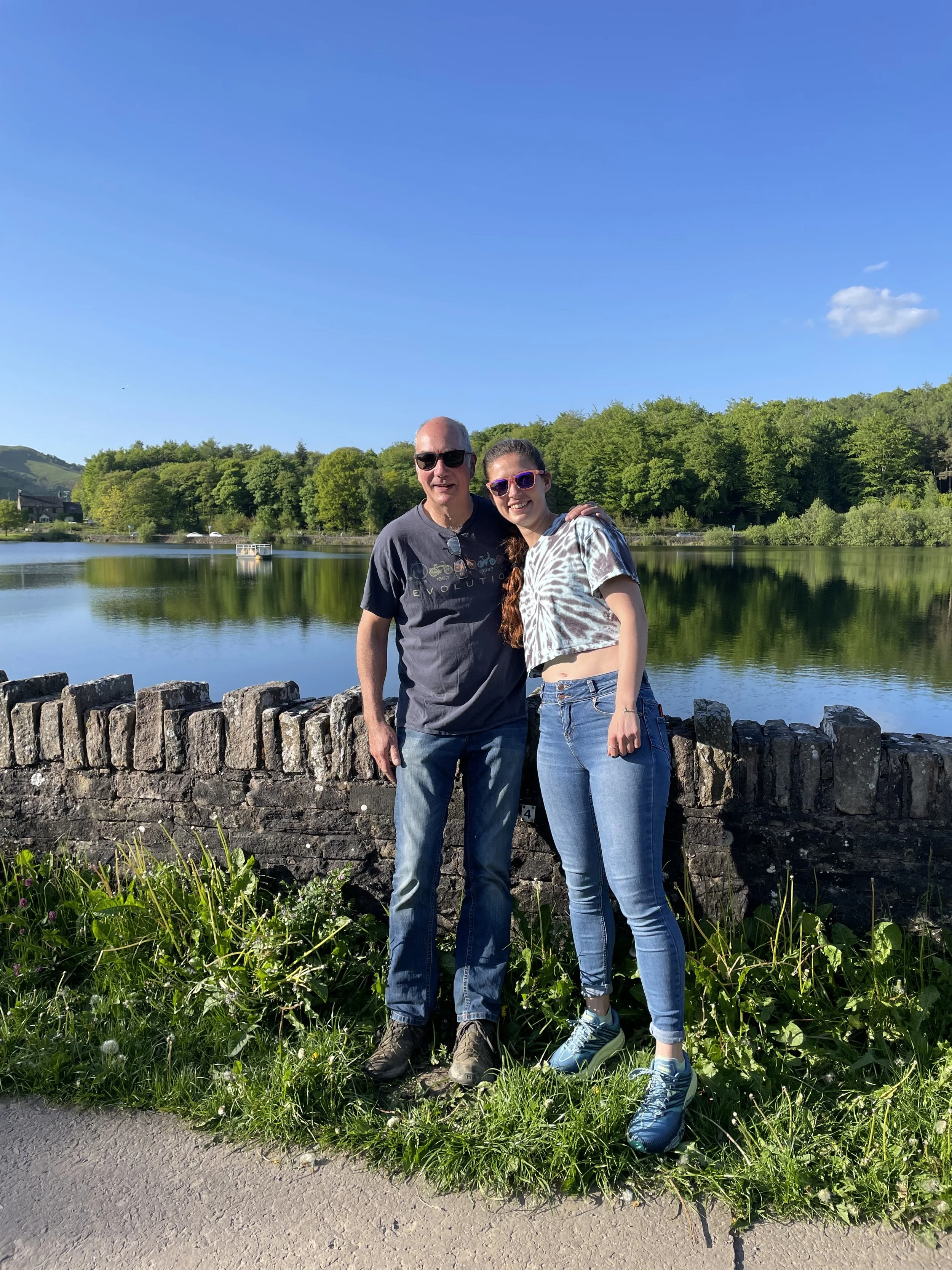 A man and woman standing outdoors by a stone wall next to a lake, smiling. The man is wearing sunglasses, a black t-shirt, jeans, and hiking shoes. The woman is wearing sunglasses, a tie-dye crop top, jeans, and sneakers. The background features a calm lake, green trees, and a clear blue sky with a few clouds.