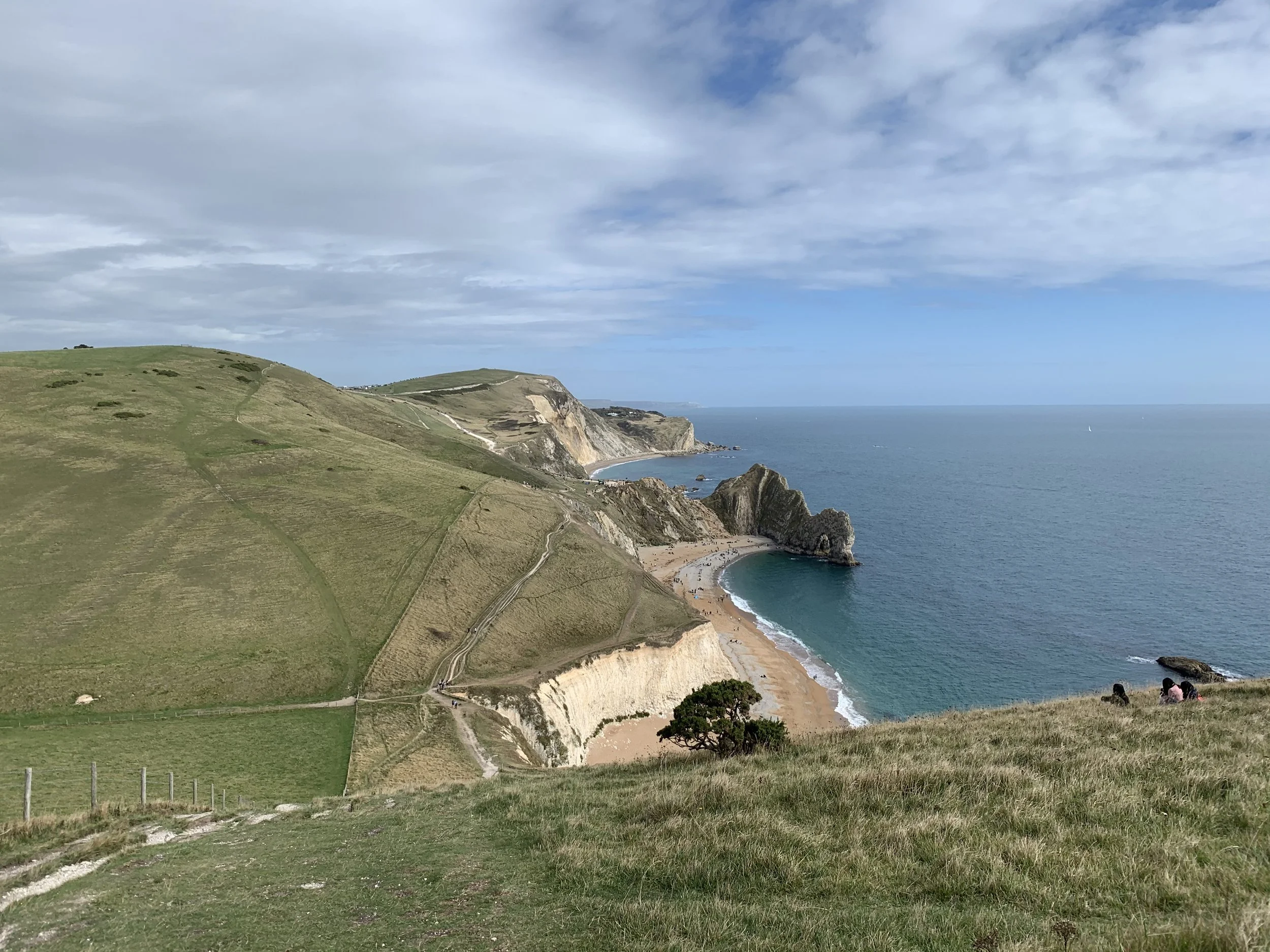 A scenic coastal landscape with grassy cliffs overlooking a beach and ocean, under a partly cloudy sky.