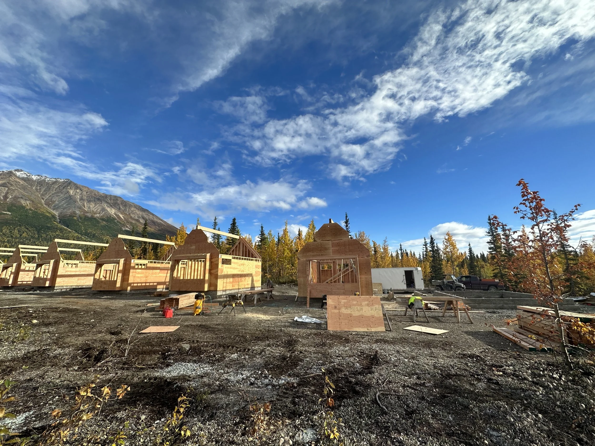 Construction site with unfinished wooden structures, tools, and equipment, set against a backdrop of mountains and a partly cloudy sky.