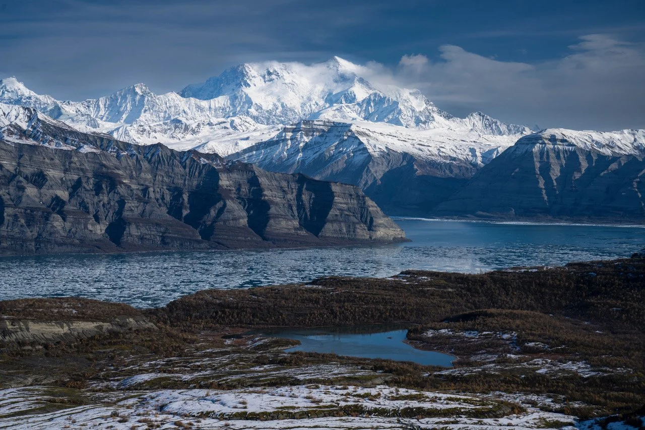 Snow-capped mountains with a large glacier, a body of water, and rugged terrain in the foreground.