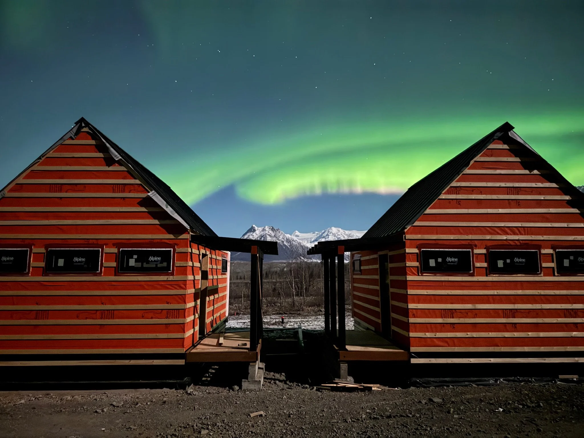 Two wooden A-frame structures with red and beige horizontal stripes, with a mountain range and aurora borealis in the night sky behind them.