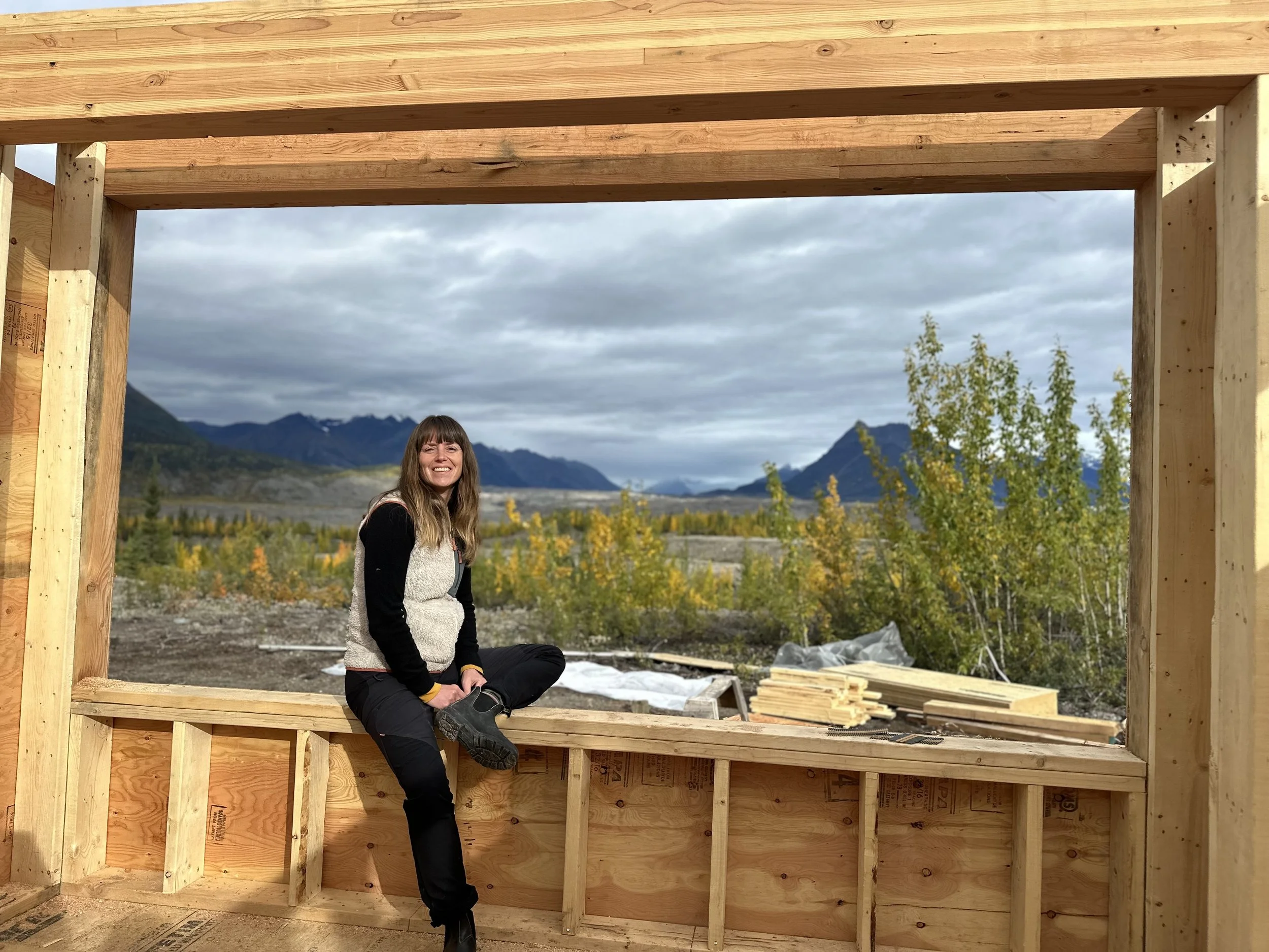 A woman smiling and sitting inside a wooden frame structure under a cloudy sky, with mountains and trees in the background during daylight.
