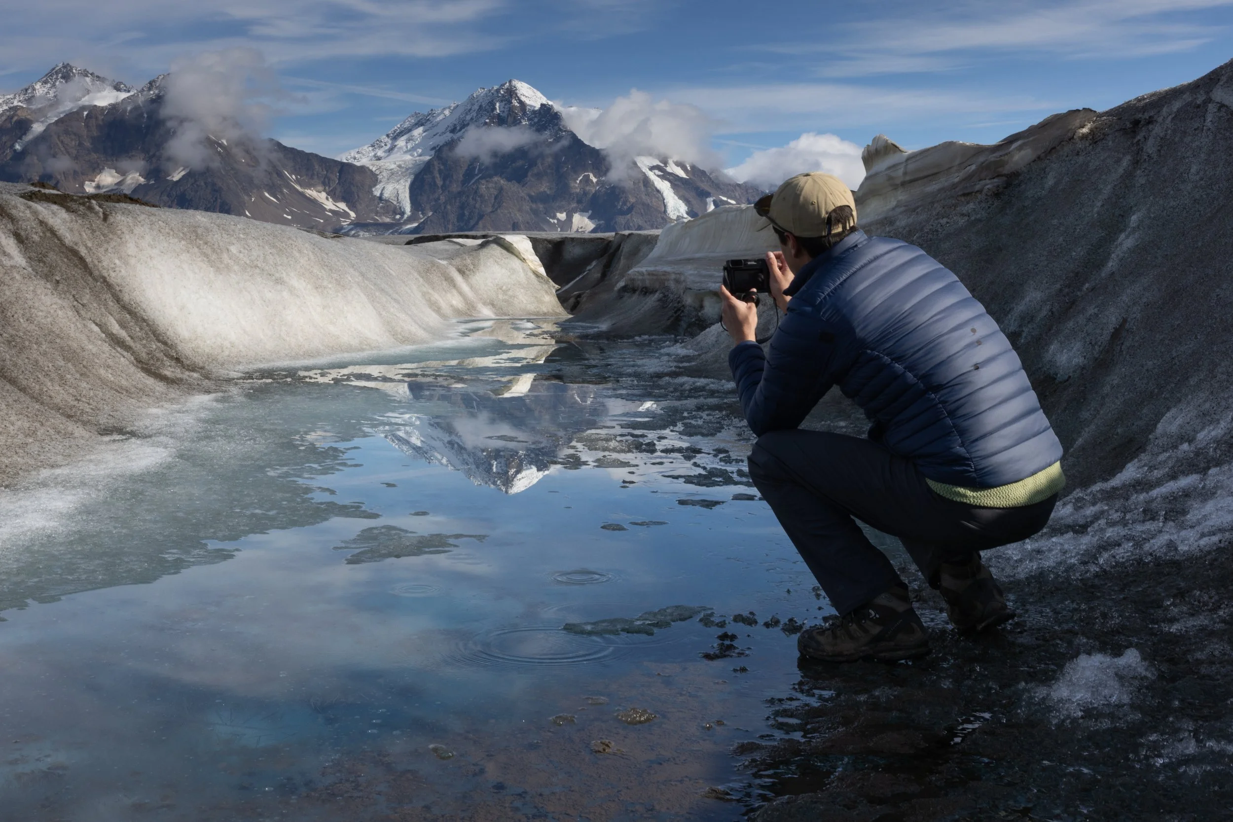 A person crouching by a small glacial meltwater stream in a mountainous landscape, with snow-capped peaks and glaciers in the background.