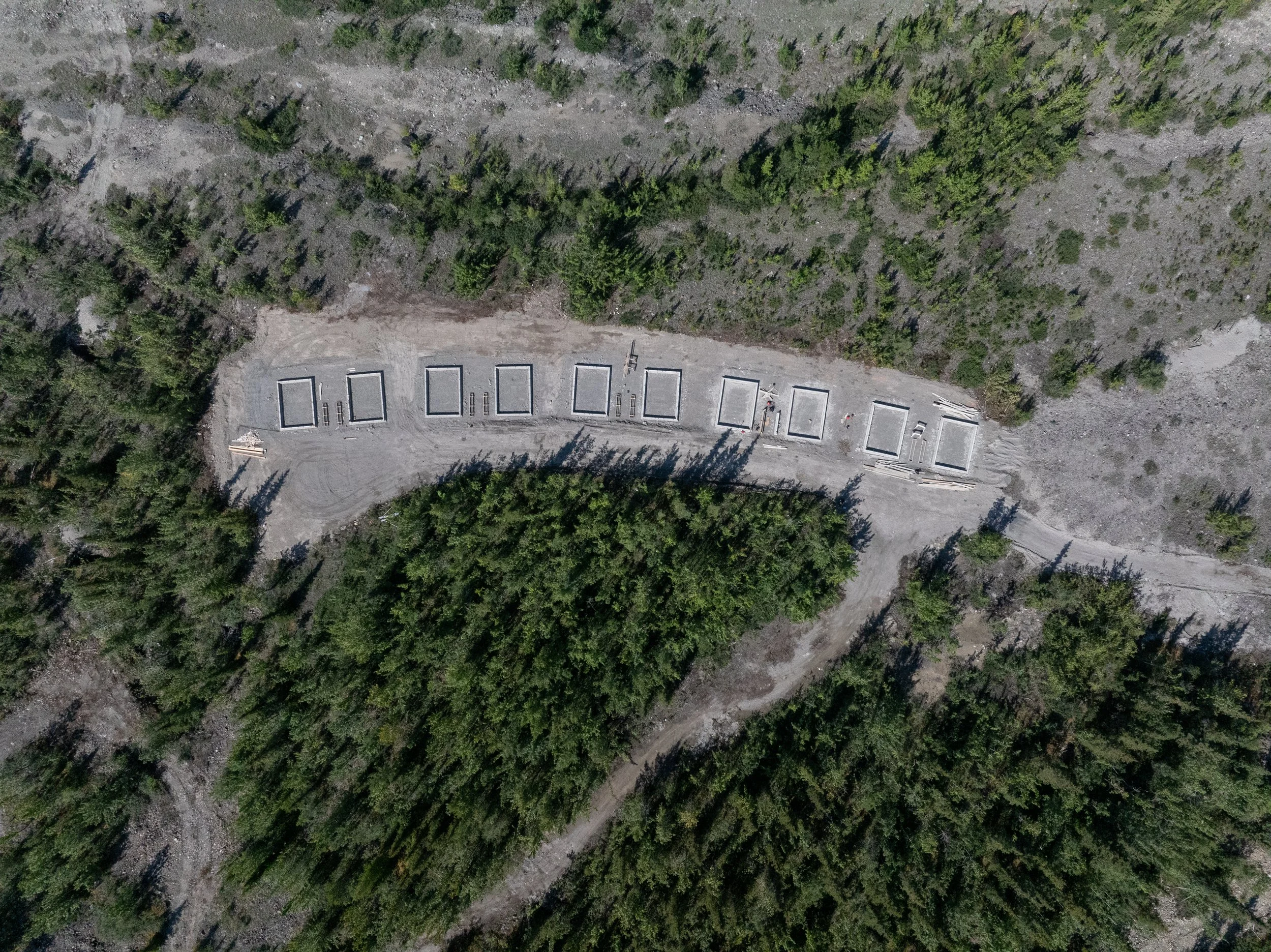 An aerial view of a forested area with a dirt construction site featuring several rectangular concrete foundations.