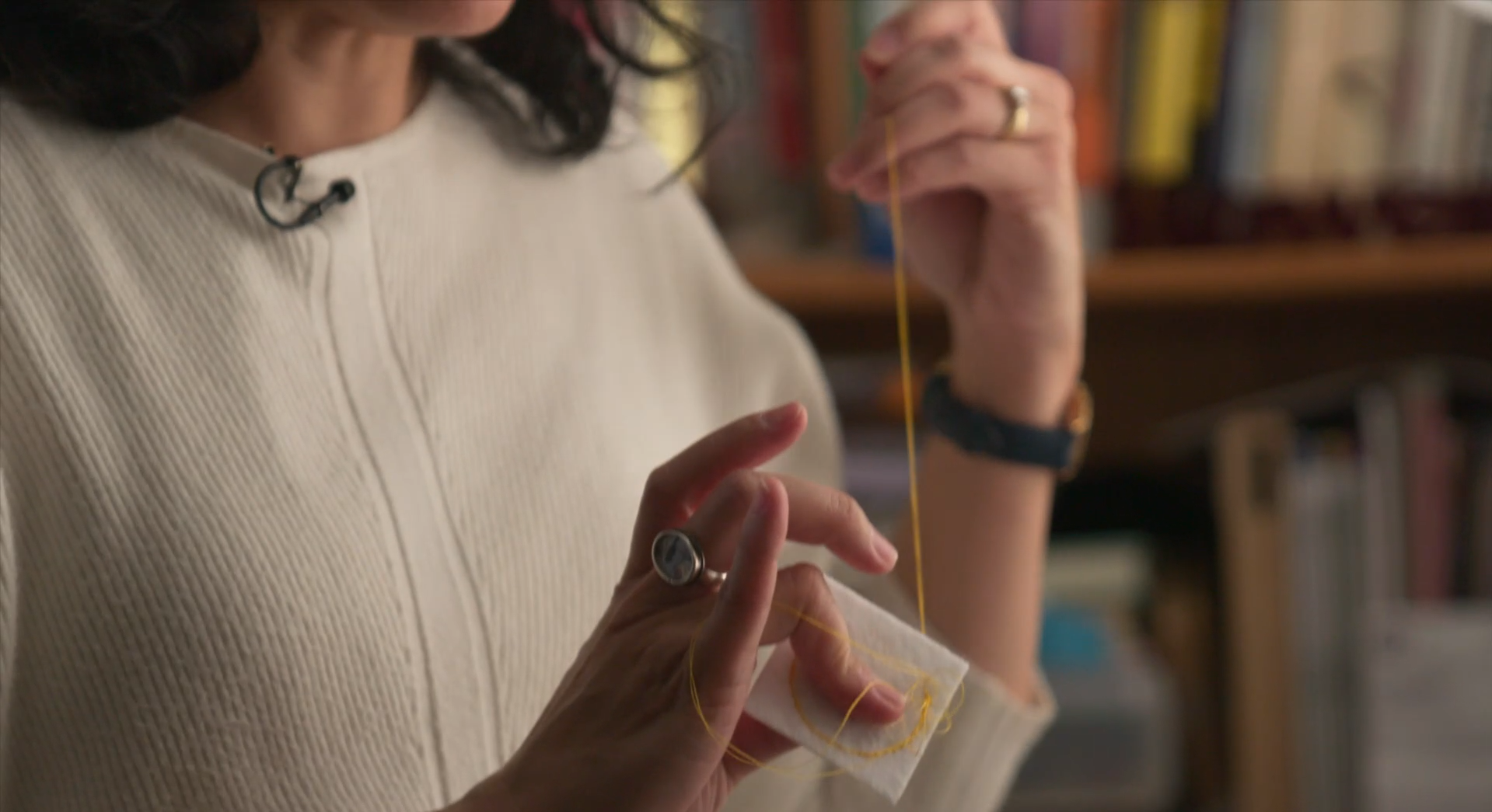 A woman with black, shoulder-length hair is hand-stitching a small square of white felt with yellow thread. The woman is wearing an ivory sweater, a ring on each hand, and a wristwatch.