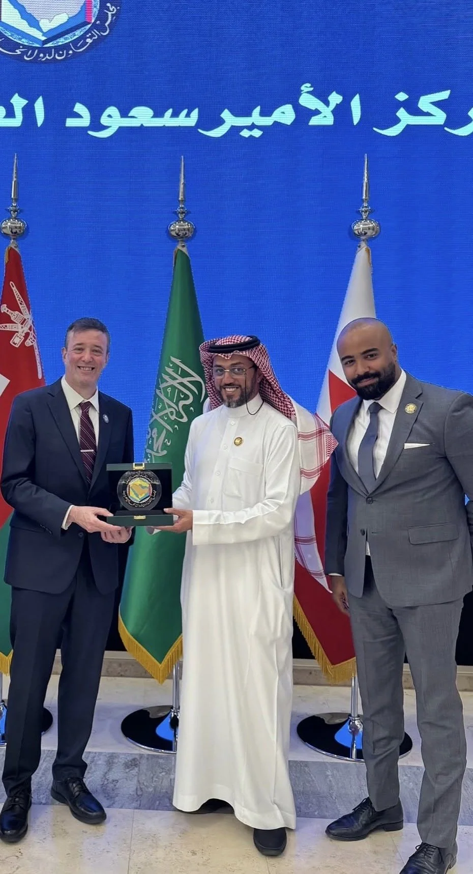 Three men in formal suits and traditional Middle Eastern attire stand in front of flags, holding a plaque together during a formal event.