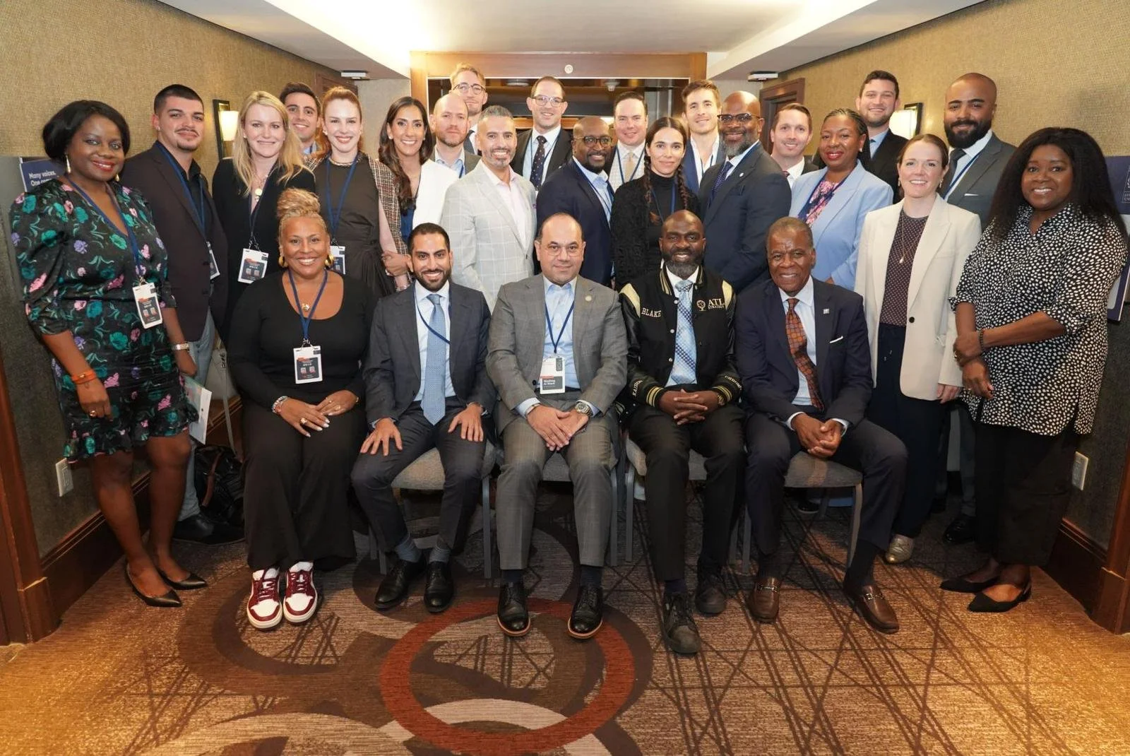 Group of professionals at a conference, posing for a photo in a hotel conference room. They are dressed in business attire and wearing conference badges.