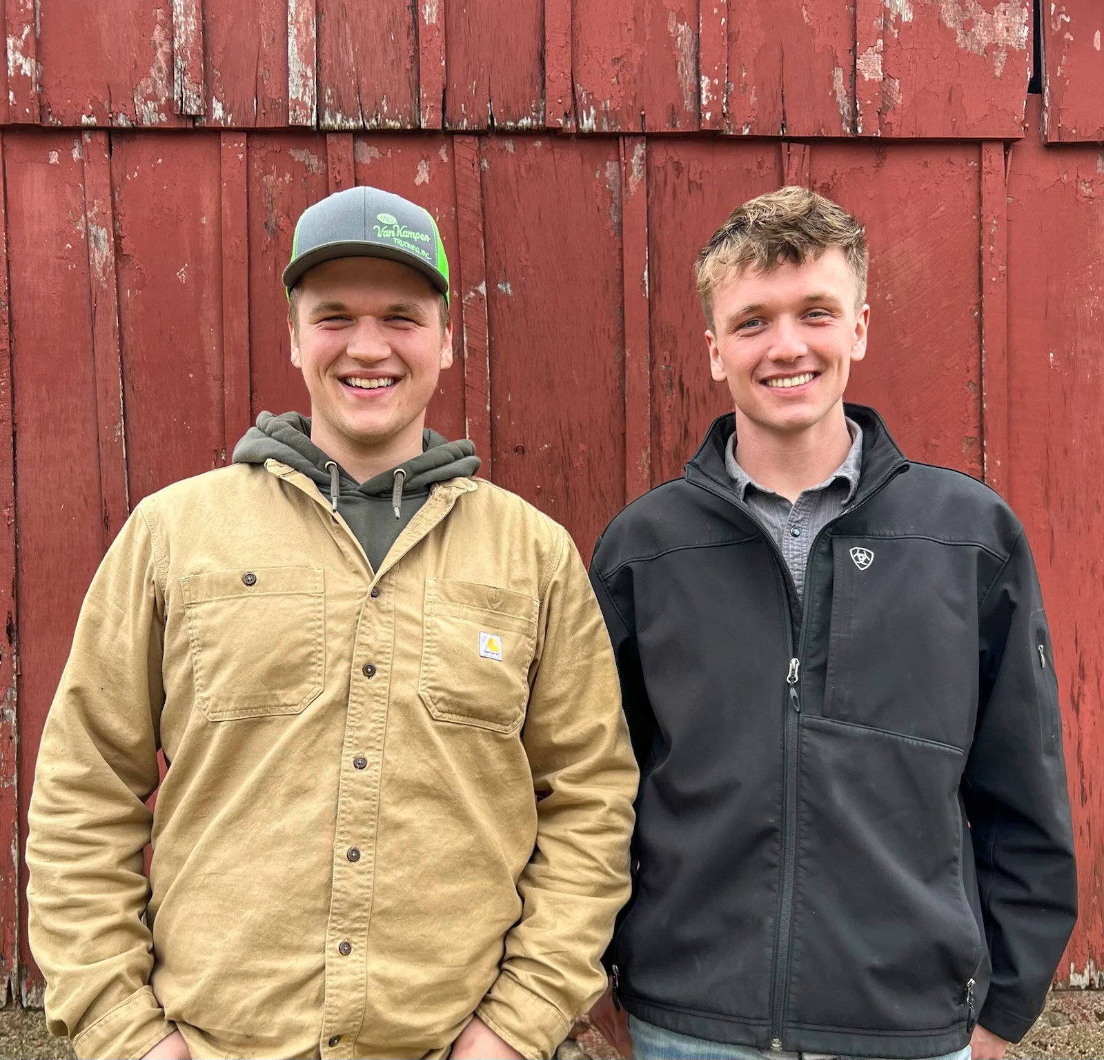 Two young men smiling, standing in front of a red wooden wall. The man on the left is wearing a gray and green baseball cap, a tan jacket over a gray hoodie. The man on the right is wearing a black jacket over a gray button-up shirt.
