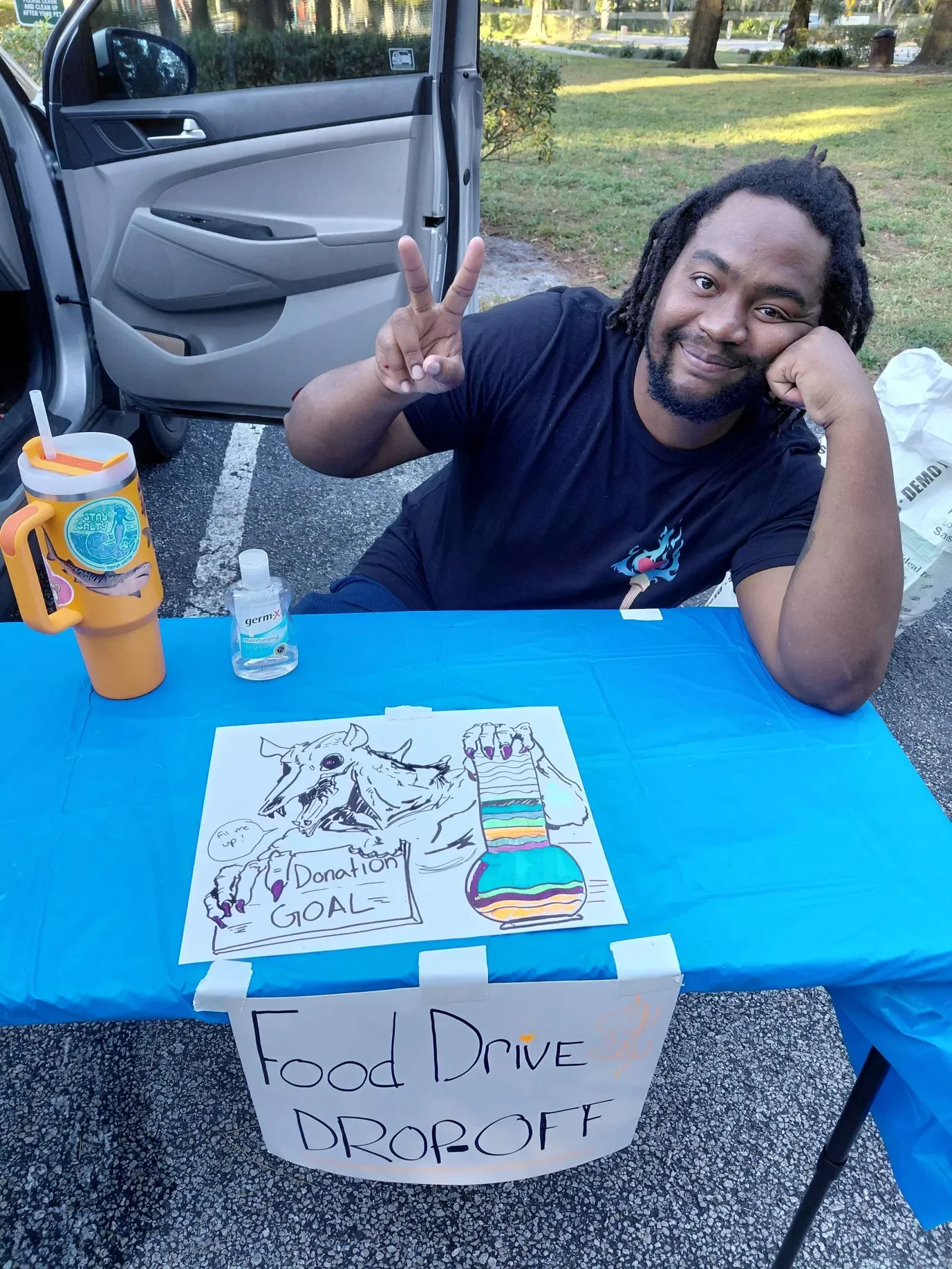 A man with dreadlocks smiling and making a peace sign with his fingers while sitting at a table outdoors. On the table, there is a coloring sheet with a skeleton and a rainbow-colored bottle, a cup with a straw, and a bottle of hand sanitizer. Behind him, a car with an open door is visible, and the scene is set in a park or green space.