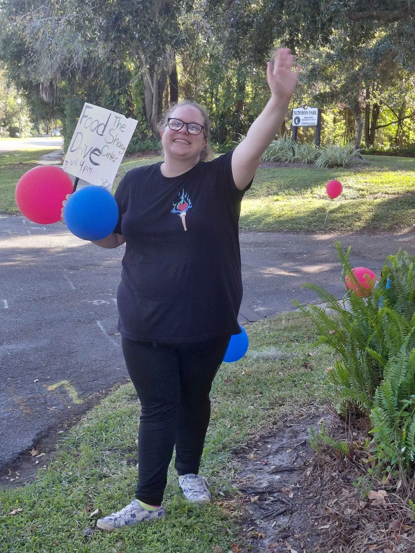 Smiling woman holding colorful balloons and a sign that reads 'The Food Share Center, 4:00 pm, Drive.' She is outdoors with trees, a sign for Audubon Park, and balloons in the background.