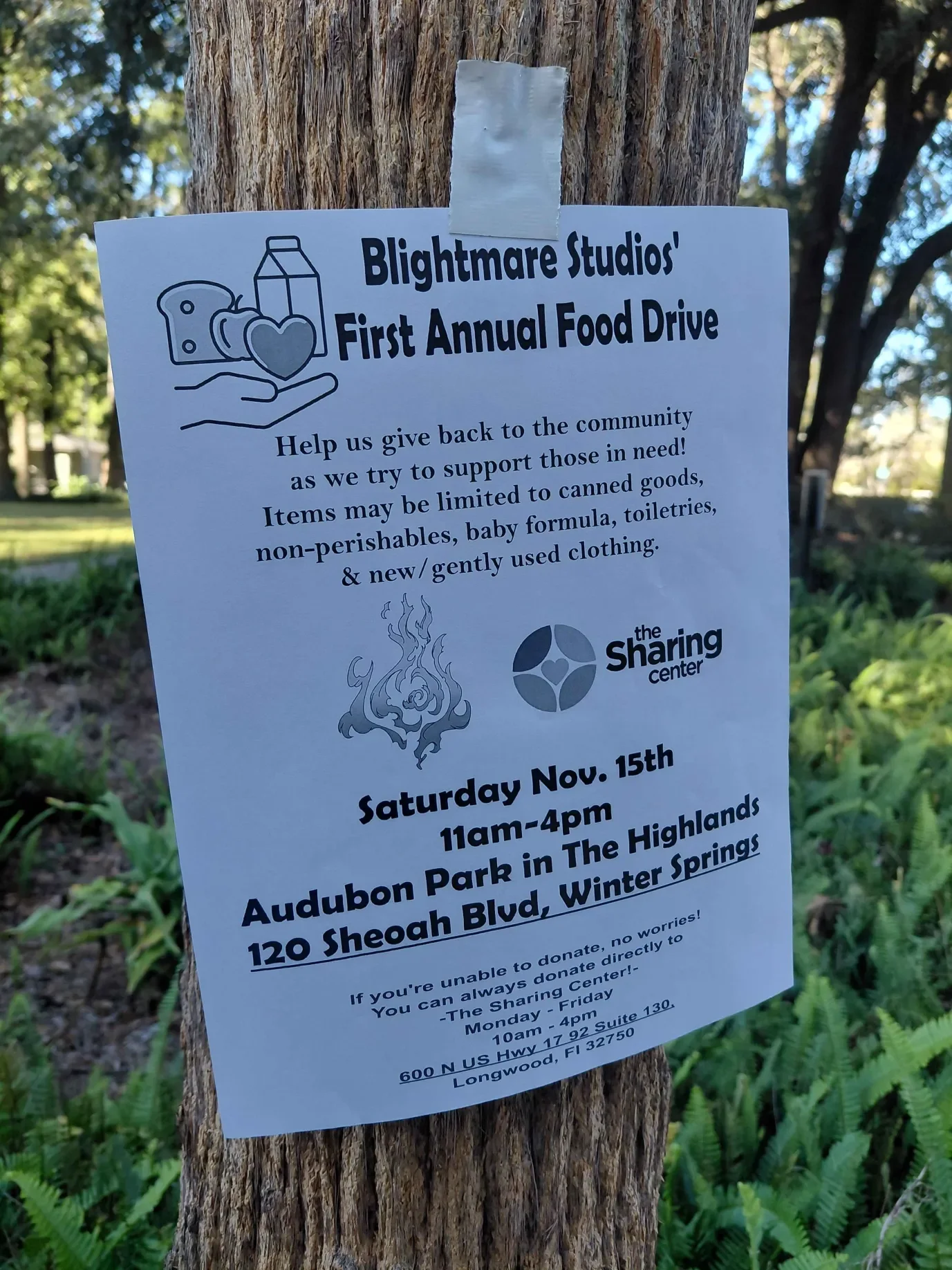 Flyer attached to a tree promoting Blightmare Studios' first annual food drive at Audubon Park in Longwood, Florida, on November 15th from 11am to 4pm. The flyer encourages donations of canned goods, non-perishables, baby formula, toiletries, and gently used clothing, with a note that items may be limited.