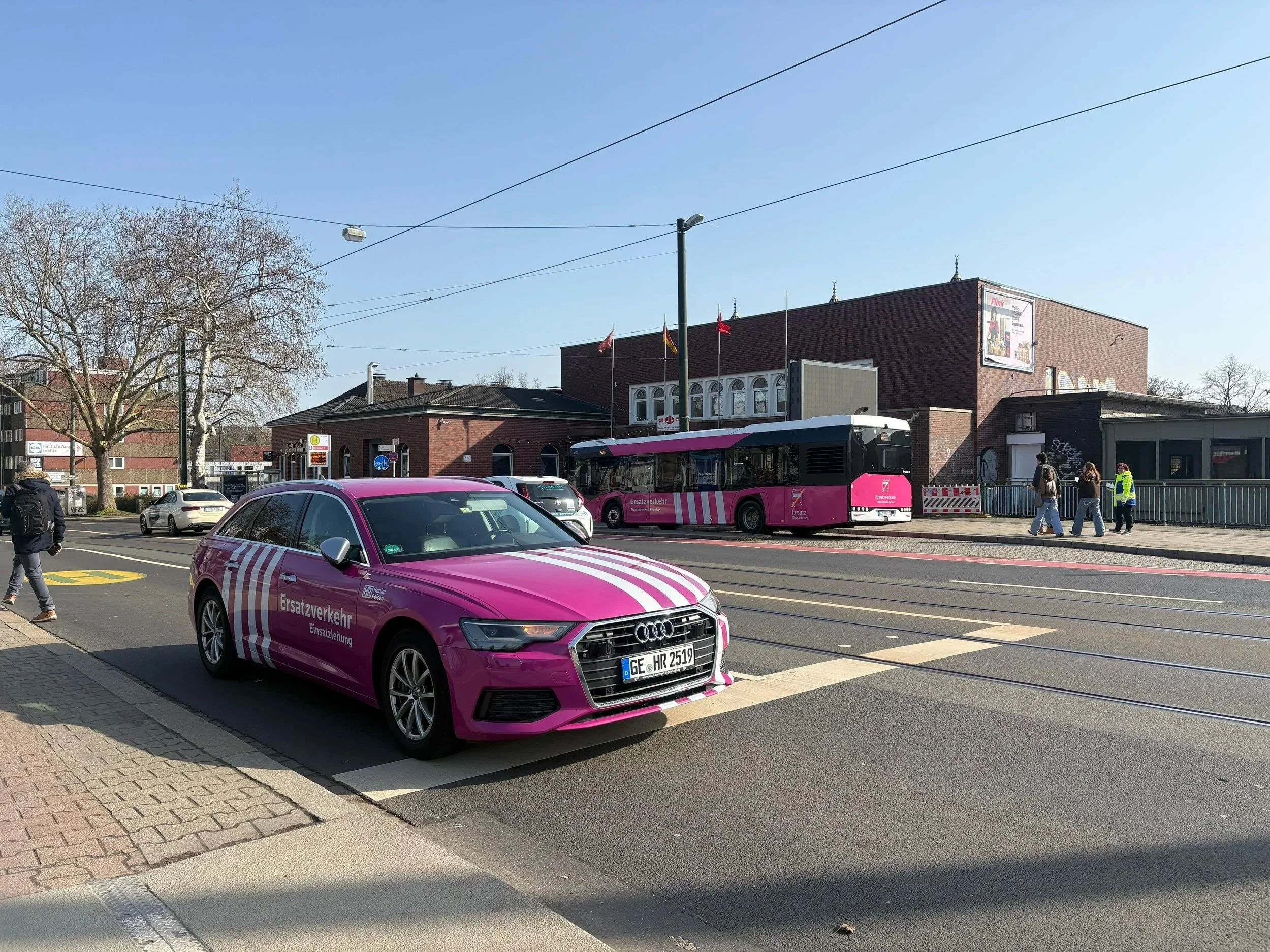 Ein pinkfarbener Audi mit der Aufschrift 'Ersatzverkehr' wird auf einer Stadtstraße gefahren, im Hintergrund eine Straßenbahn und einige Fußgänger.