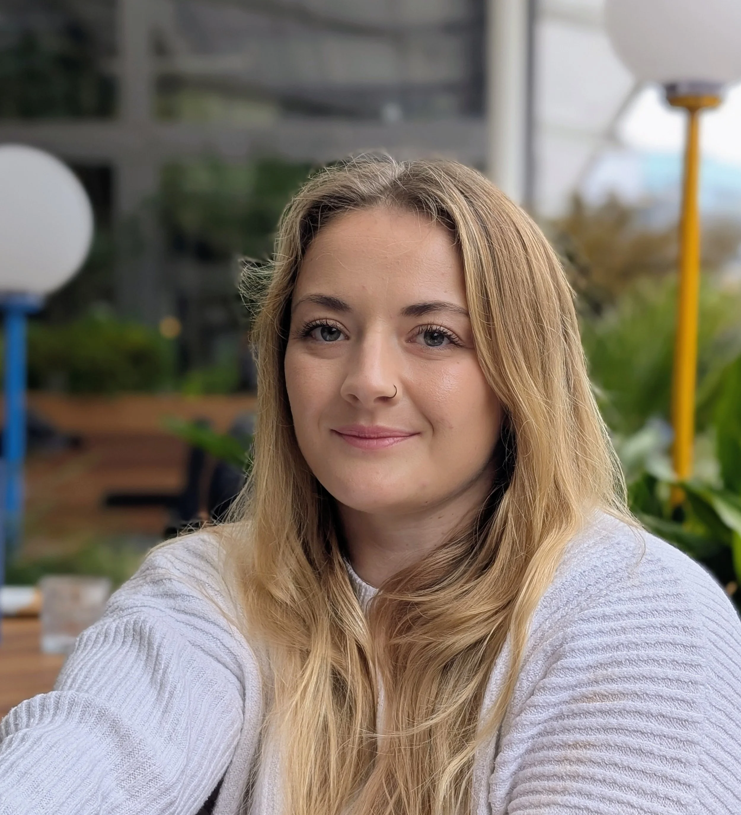 A woman with long blonde hair, fair skin, and a nose piercing smiling at the camera, seated outdoors with green plants and outdoor furniture in the background.
