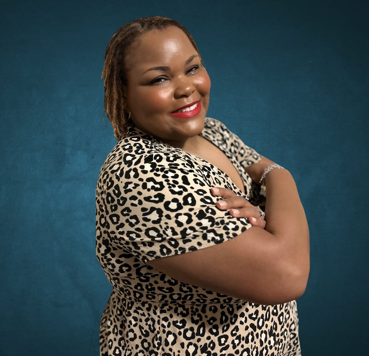 A woman with short dreadlocks, smiling with red lipstick, wearing a leopard print dress, standing against a solid teal background.