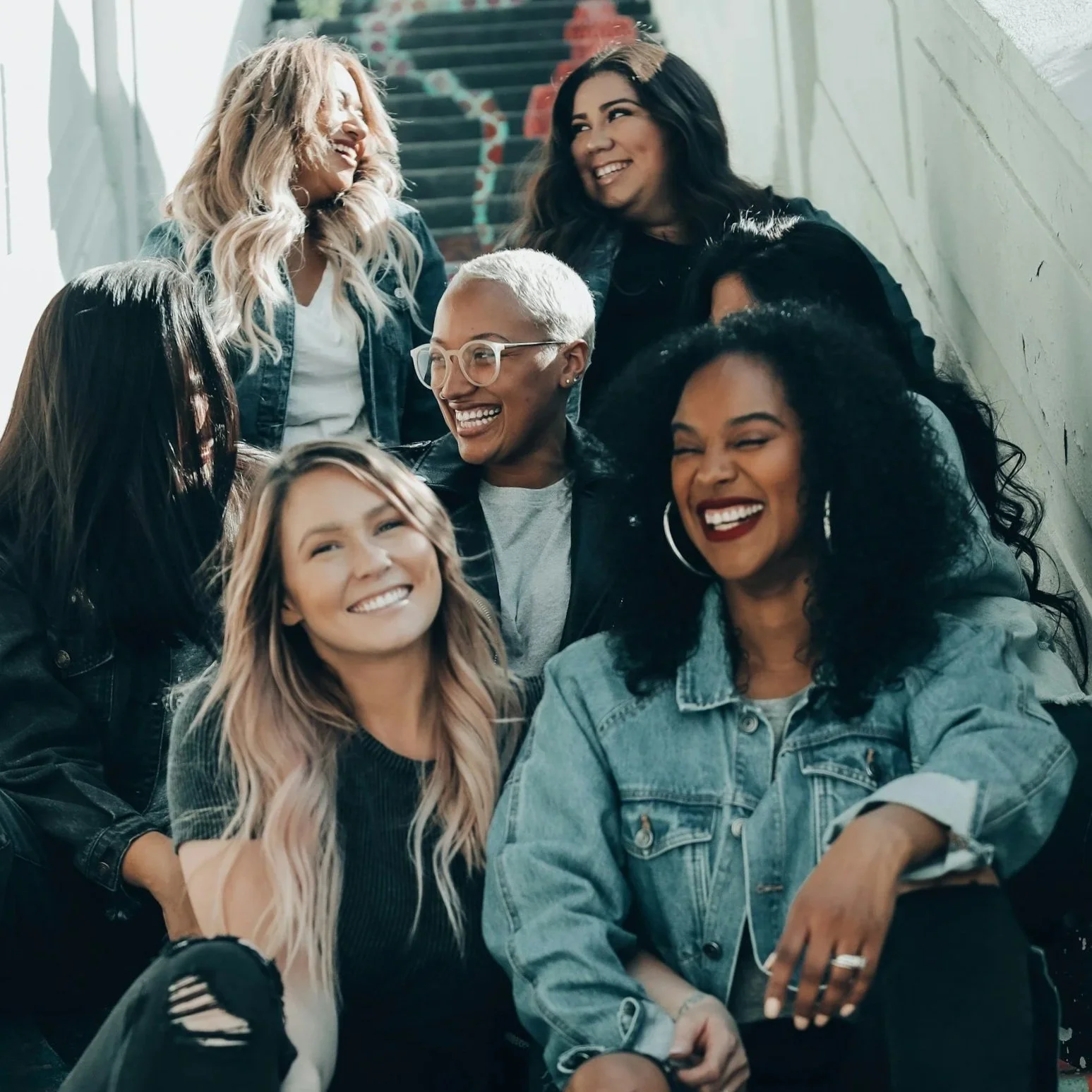 A group of six women of diverse ethnicities sitting and standing on a staircase, smiling and enjoying each other's company in a well-lit indoor space.