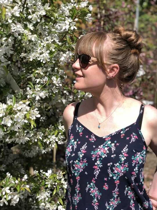 Side view of a woman with sunglasses, floral dress, and a bun hairstyle, standing near white flowering bushes in sunlight.