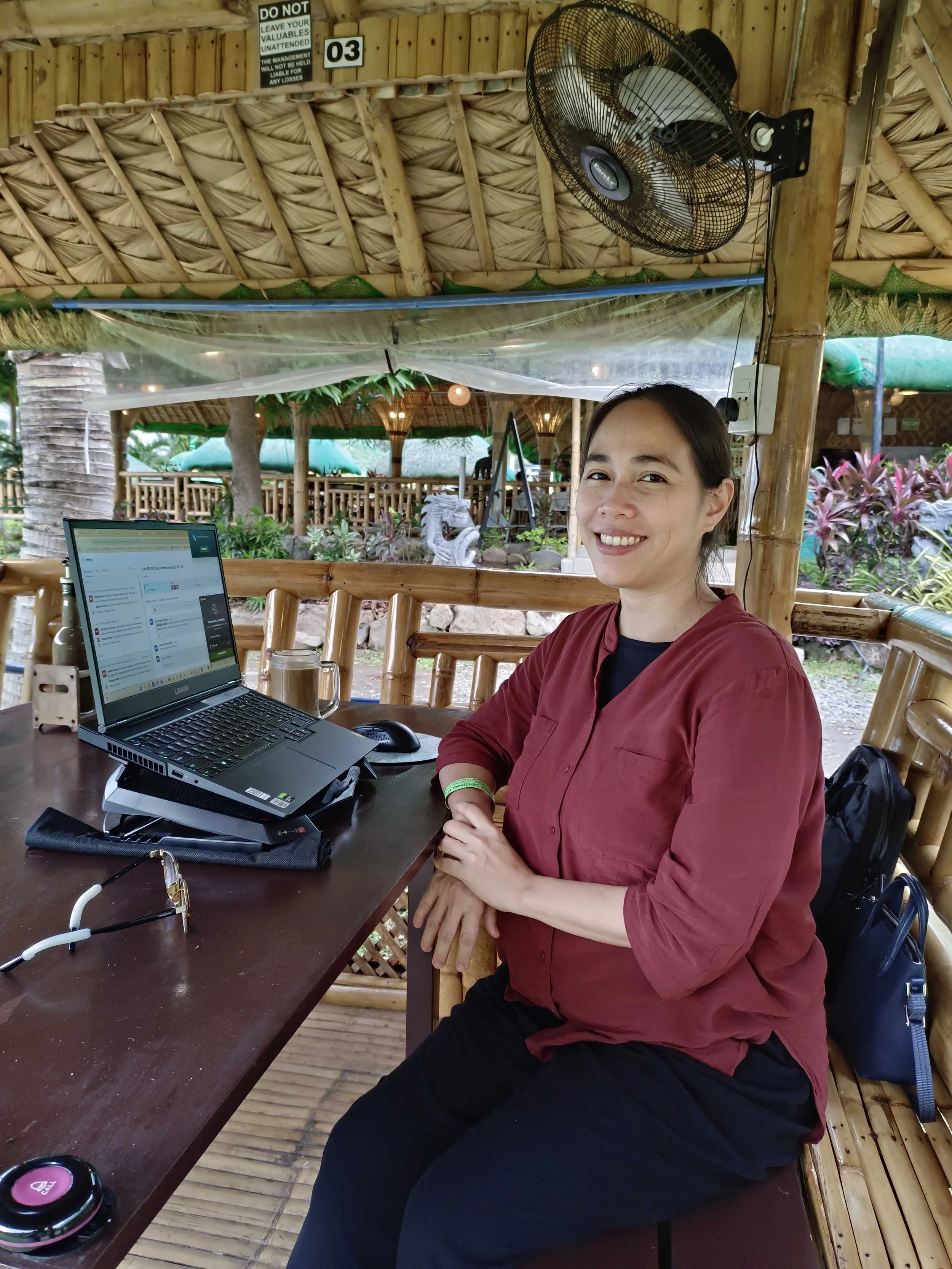 Smiling woman sitting at a wooden table in an outdoor bamboo and thatch setting with a laptop, sunglasses, and a drink, surrounded by tropical plants.