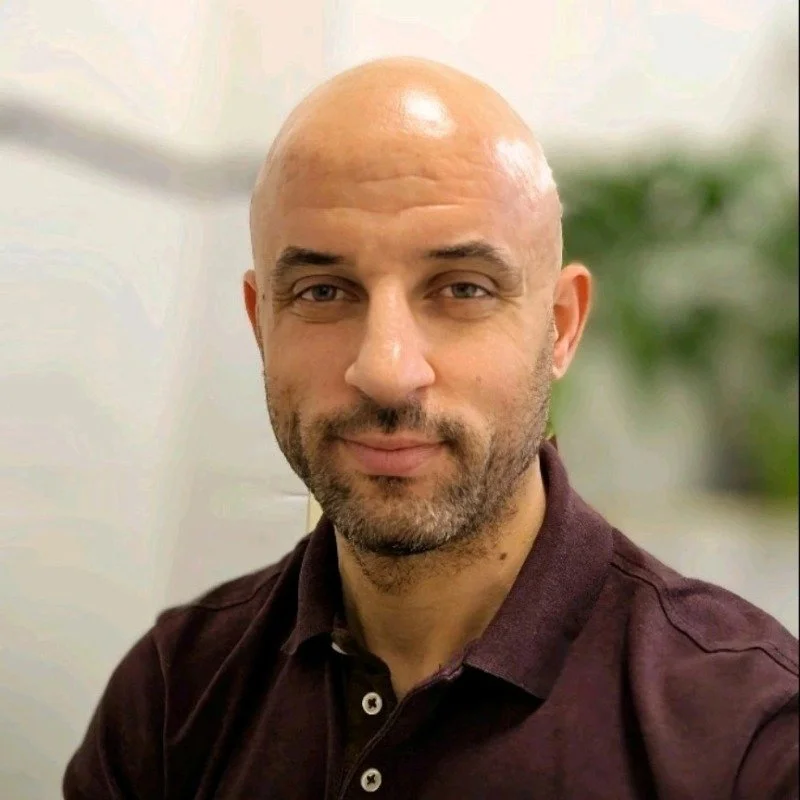 A bald man with a beard and mustache, wearing a dark maroon shirt, poses for a selfie in an indoor setting with a blurred background and some greenery.