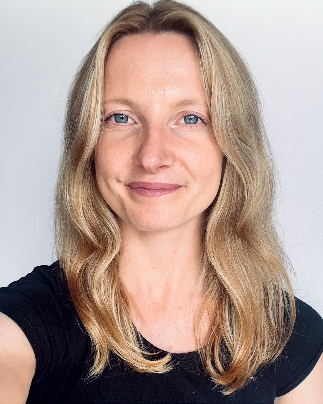 A woman with long blonde hair and blue eyes smiling, wearing a black shirt against a plain white background.