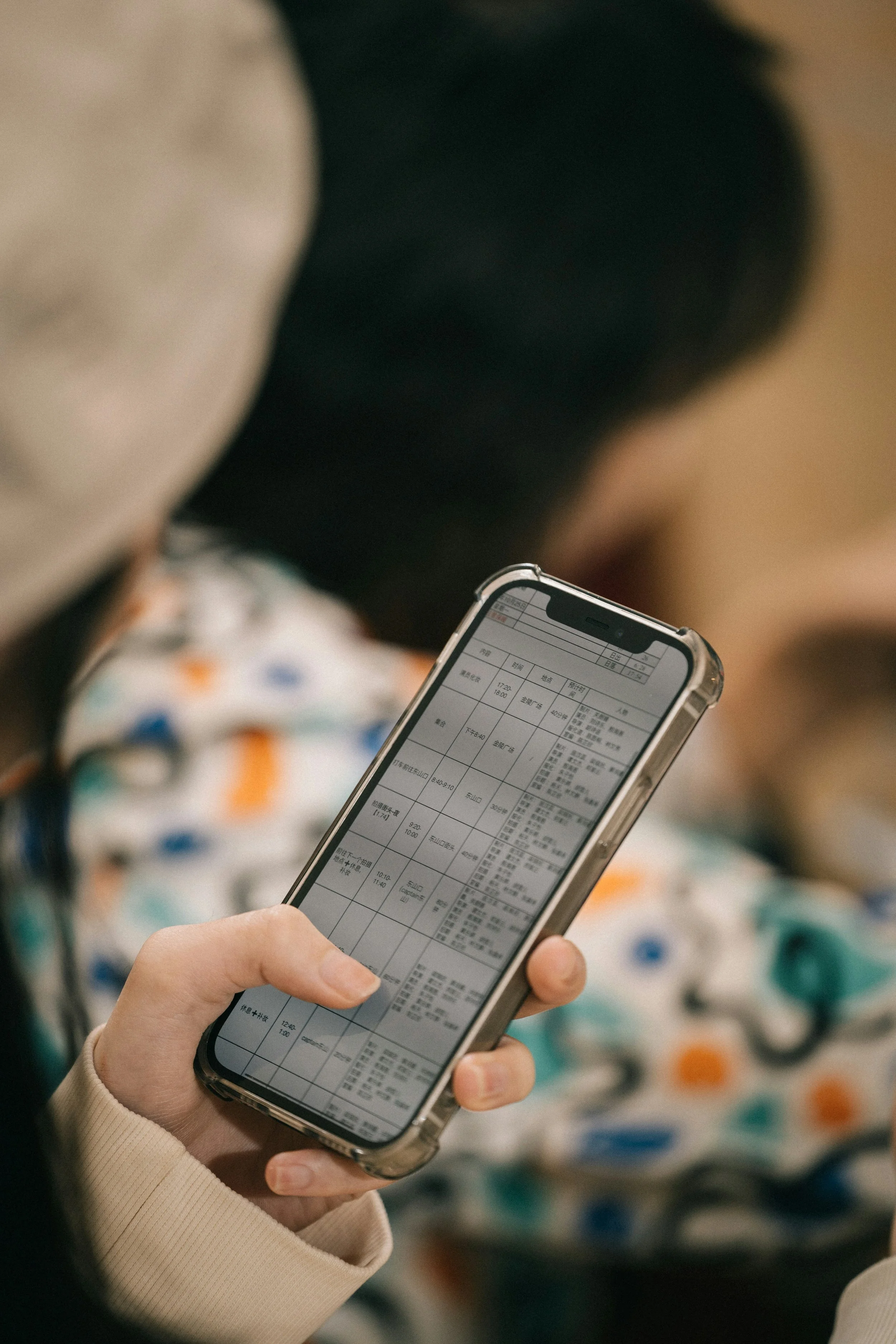Close-up of a person using a smartphone to view a schedule or timetable, with a blurred background showing a person in a colorful, patterned shirt.
