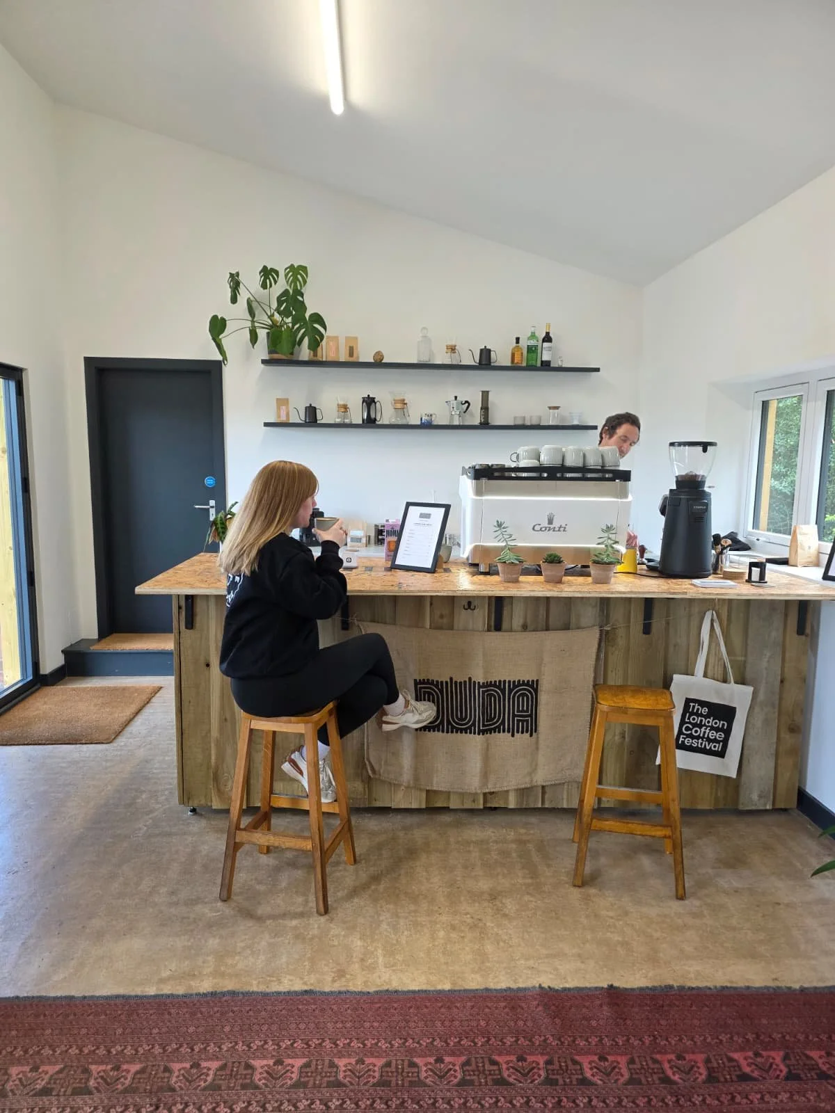 A woman sitting on a stool at a wooden coffee bar, sipping a drink, with a barista preparing coffee behind the counter in a coffee shop with white walls, black shelves, and green plants.