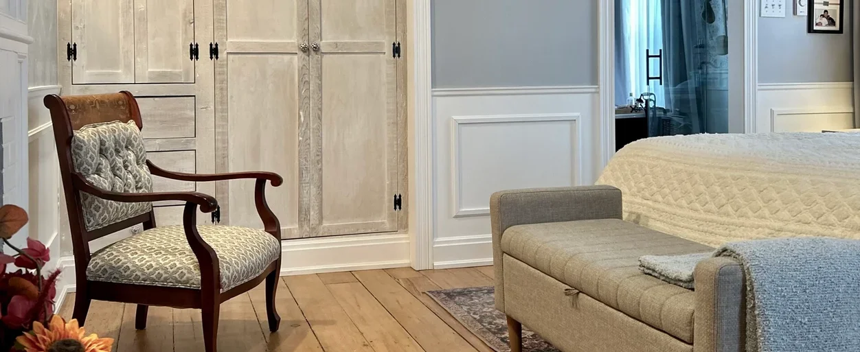 A cozy bedroom corner with a vintage wooden armchair with patterned upholstery, a beige upholstered bench, and a bed with a cream-colored quilt. There are built-in cabinets and a window with light curtains.