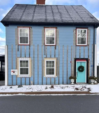 Blue two-story house with four windows, a turquoise door with a wreath, a small potted plant, and a wooden fence in front, with snow on the ground.