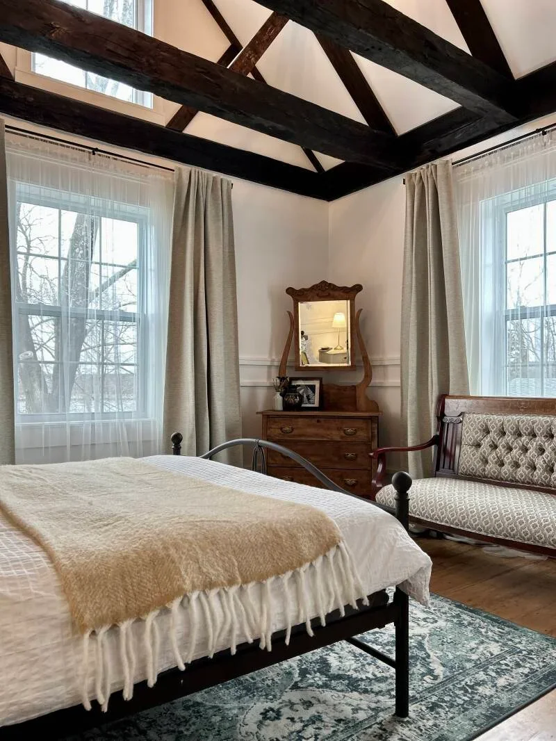 A cozy bedroom with a black metal bed, beige blanket, and wooden dresser with a mirror. Two large windows with sheer white curtains and beige drapes, wooden floor, vintage sofa, and exposed dark wooden beams on the ceiling.