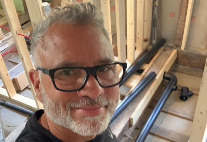 Man with glasses and gray beard smiling inside a construction site with wooden framing and pipes.