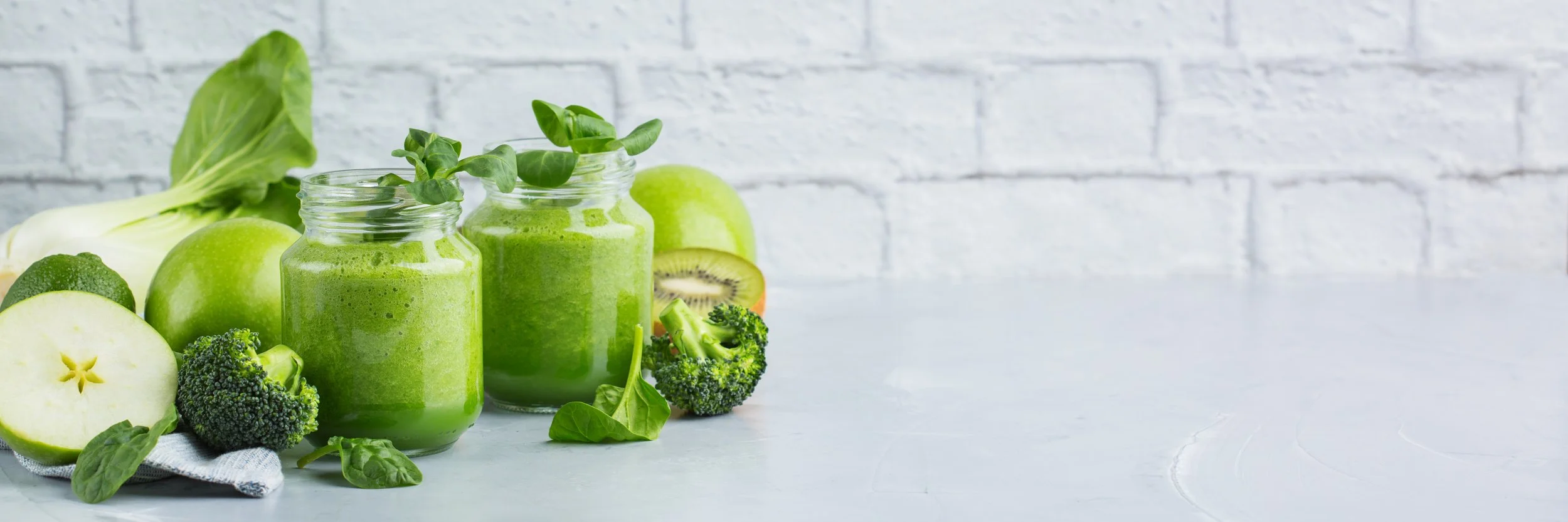 Green smoothies in glass jars surrounded by green apples, broccoli, spinach leaves, and kiwi on a white surface with a white brick wall background.