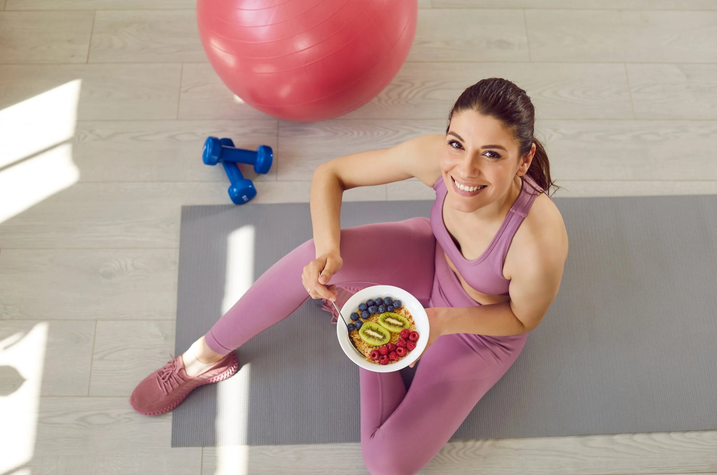 A woman in pink workout clothes sitting on a yoga mat, holding a bowl of fruit and smiling at the camera. There is a large pink exercise ball and a pair of blue dumbbells nearby.
