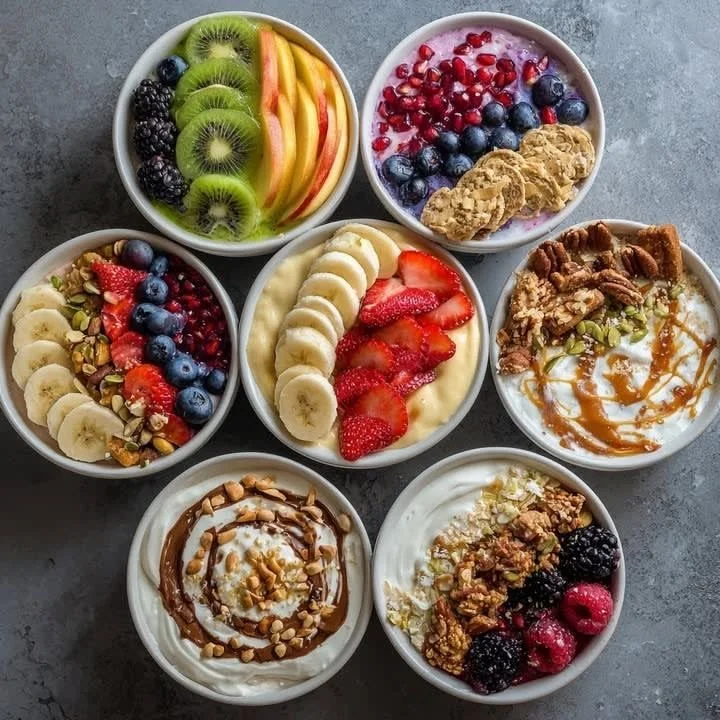 Seven bowls of yogurt topped with various fruits, granola, and drizzles, including kiwi, apple, blackberry, blueberry, pomegranate, banana, strawberry, raspberry, nuts, caramel, and chocolate.