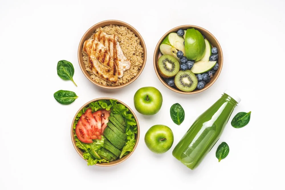 Bowls of grilled chicken with quinoa, mixed salad with tomato and avocado, and a fruit salad with kiwi, apple, and blueberries, along with two green apples, spinach leaves, and a green smoothie bottle on a white background.