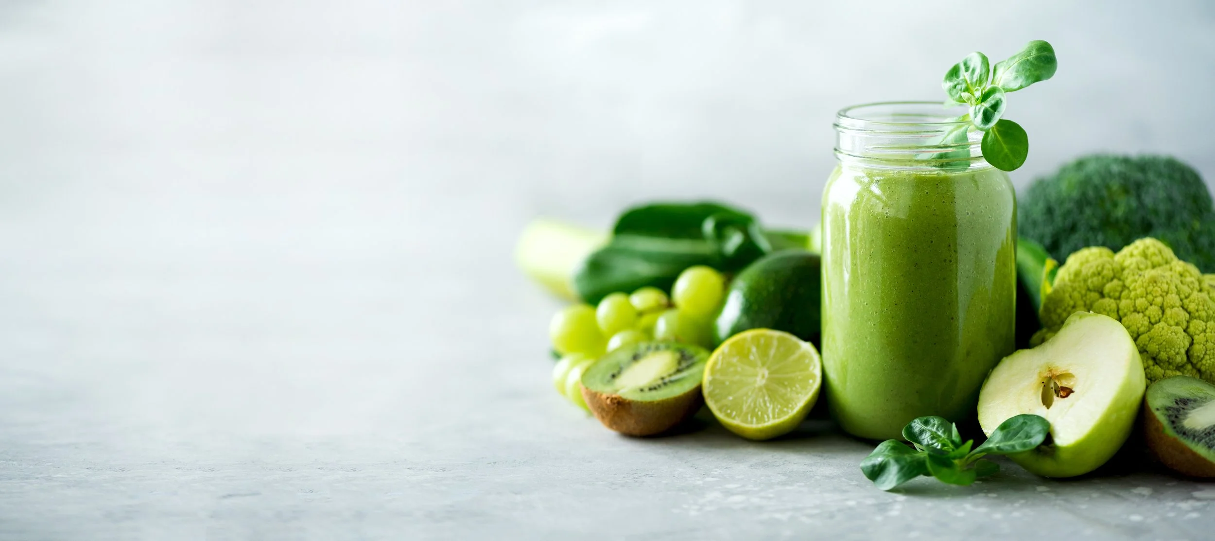 A green smoothie in a glass jar garnished with a small green leafy herb, surrounded by green fruits and vegetables including kiwi, lime, cucumber, broccoli, and green grapes on a white surface.