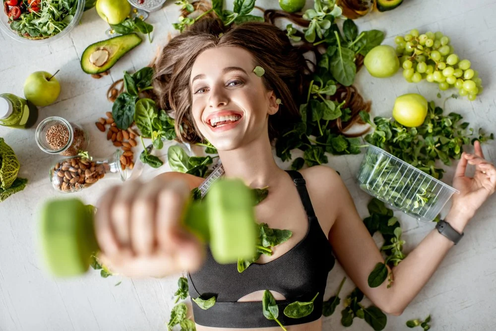 A smiling woman lying on the floor surrounded by green apples, avocados, leafy greens, nuts, and salad ingredients, holding a green dumbbell in one hand.
