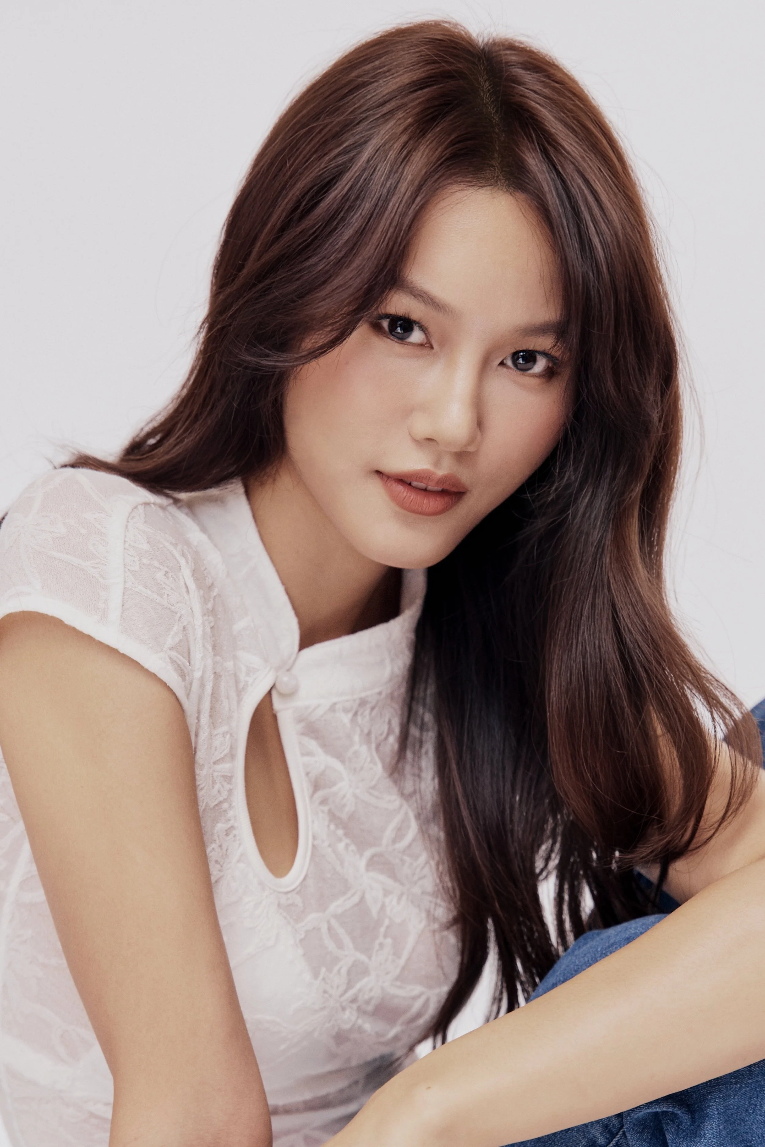 Close-up portrait of a young woman with long brown hair, wearing a white lace top, looking directly at the camera.