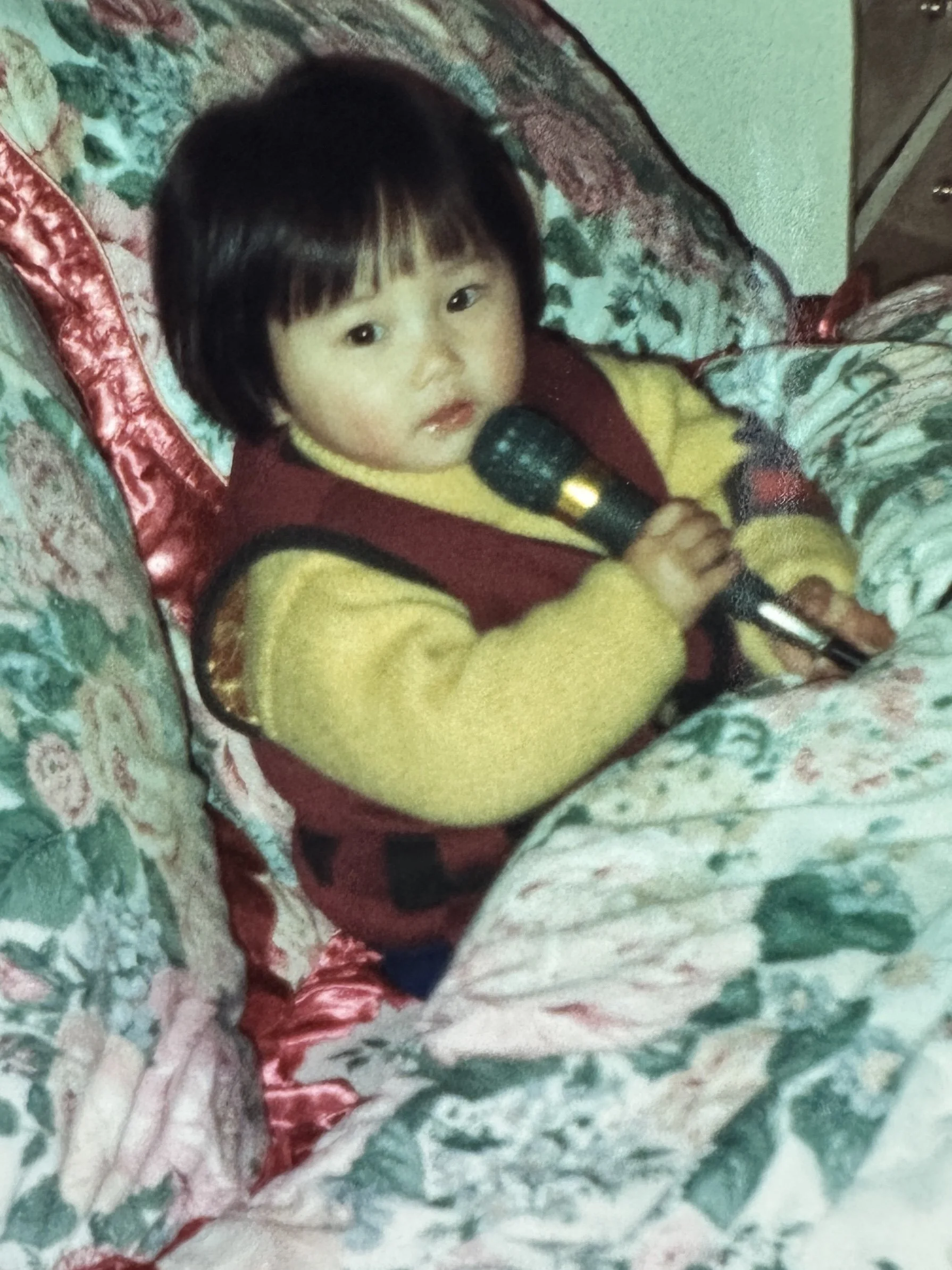 A young child with dark hair and bangs, wearing a yellow sweater and a red vest, sitting on a floral-patterned couch and holding a microphone.