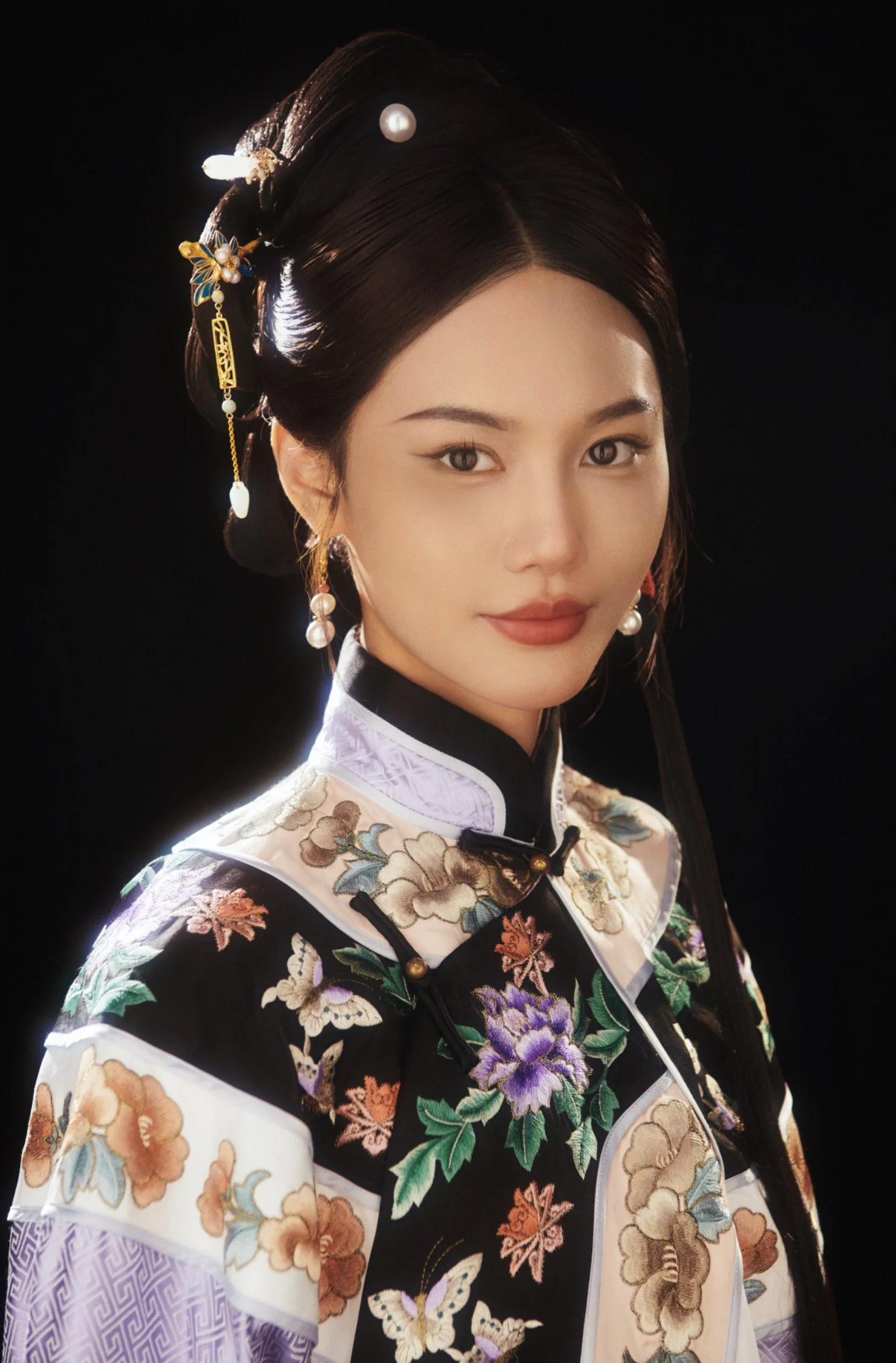 Young woman in traditional Asian attire with floral and butterfly embroidery, wearing ornate hairpieces and pearl earrings, against a black background.