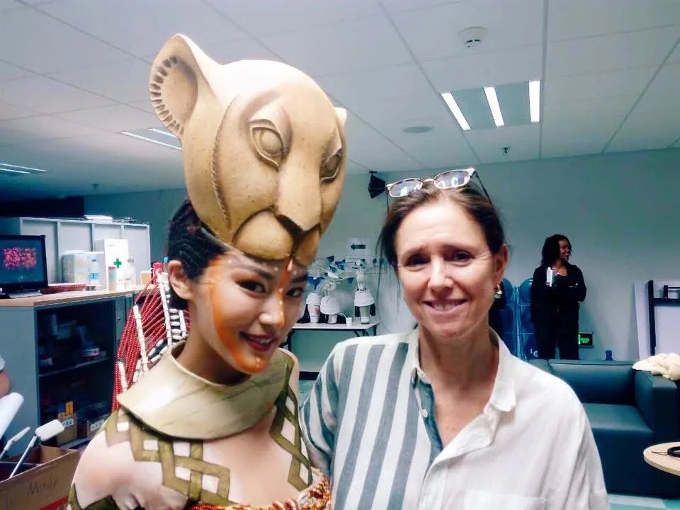 A performer in colorful costume and face paint stands next to a woman in a white striped shirt, both smiling in an indoor setting.