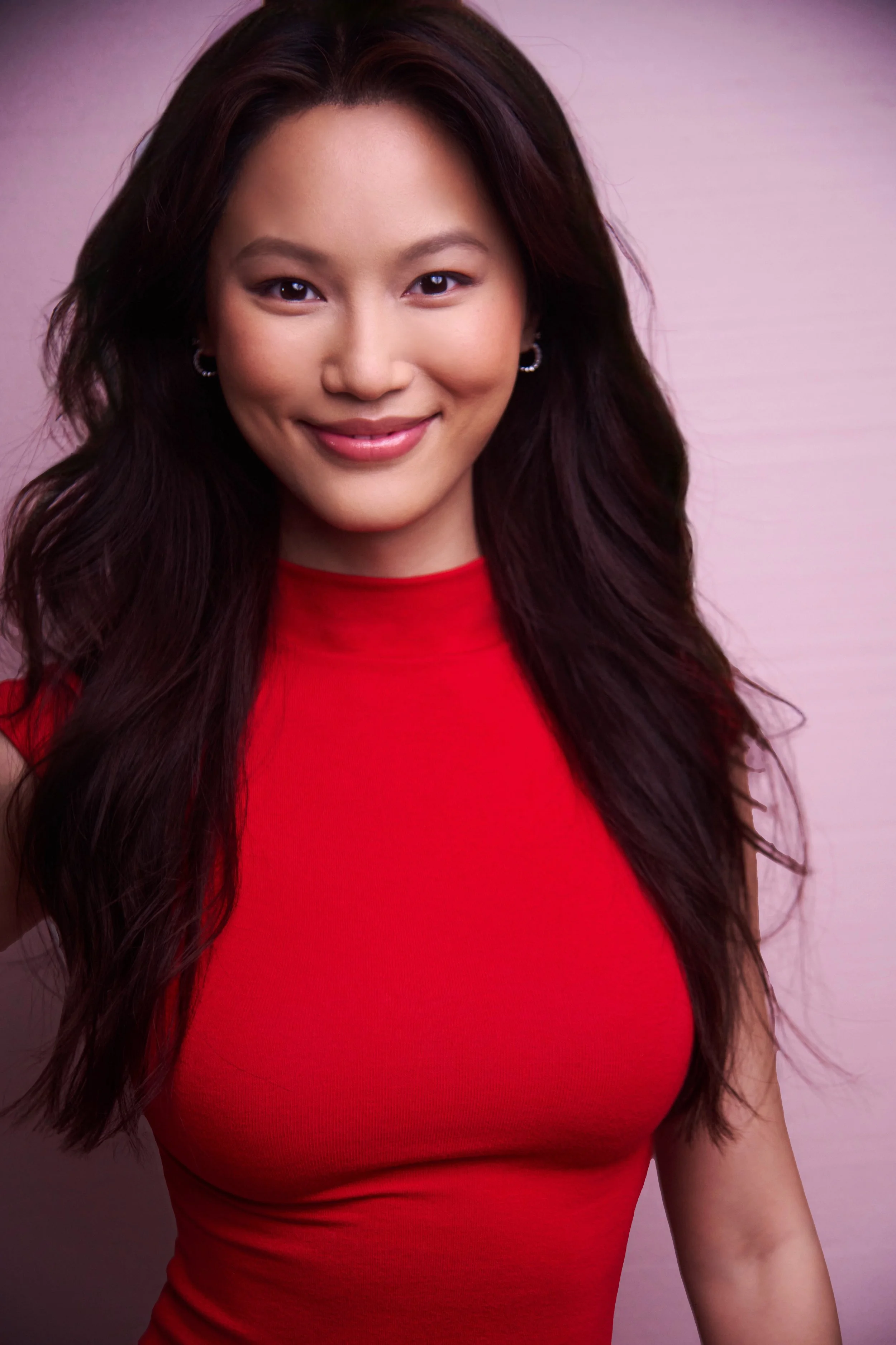 Portrait of a young woman with long dark hair, wearing a red top, smiling, against a light pink background.