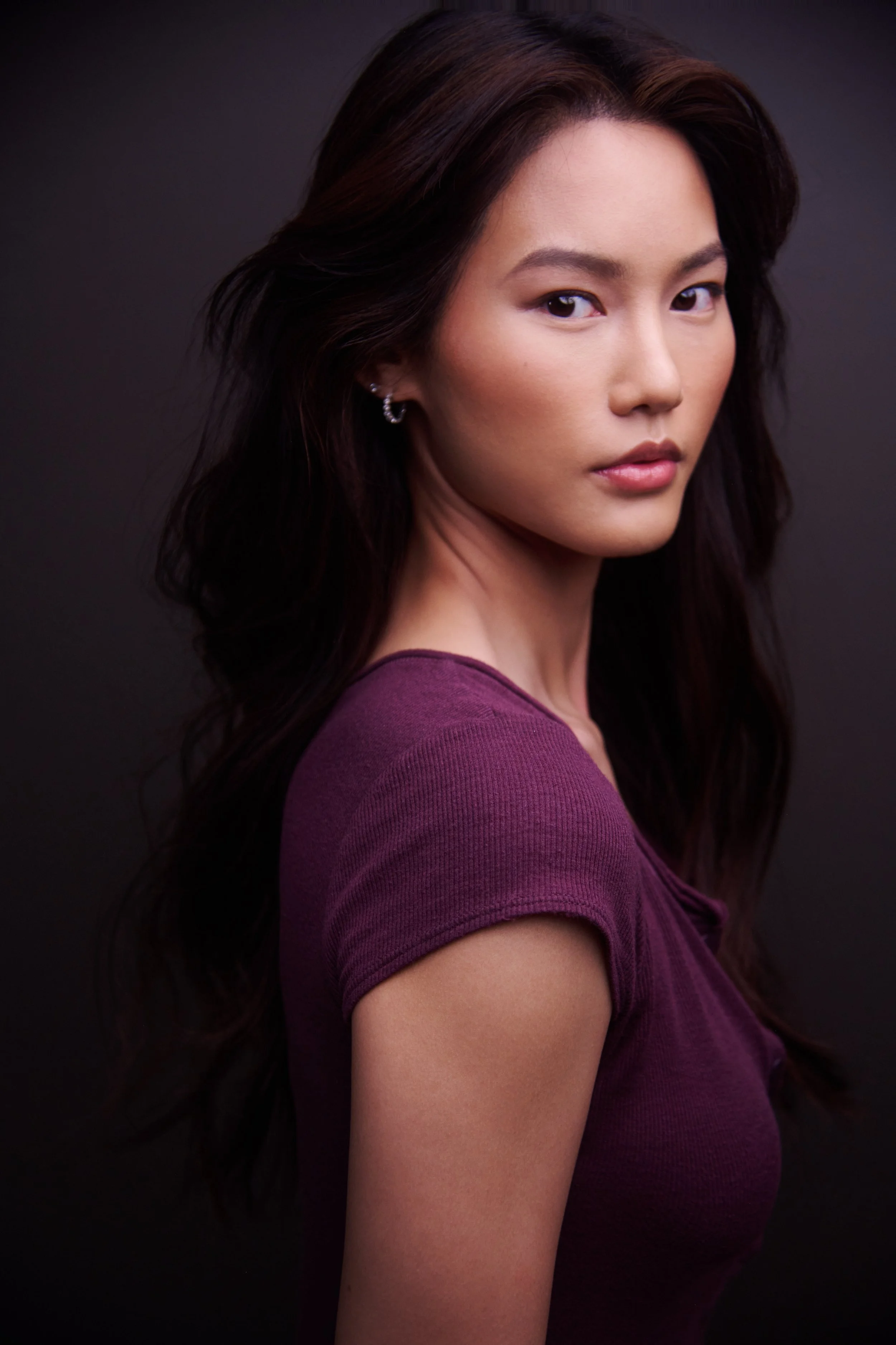 Portrait of a young woman with long dark hair, wearing a purple top, against a dark background.