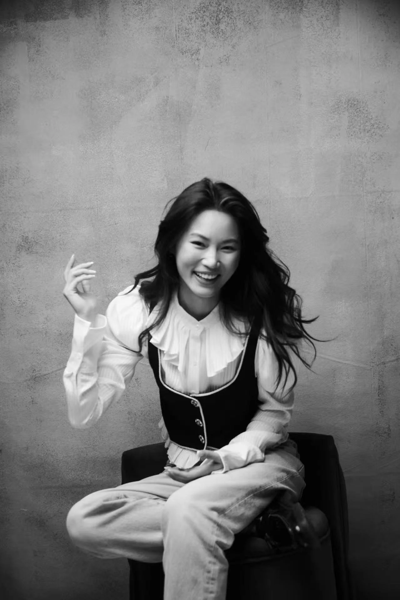 Black and white photo of a smiling woman with long wavy hair sitting on a small stool against a plain wall. She is wearing a ruffled blouse, a vest, and light-colored pants.